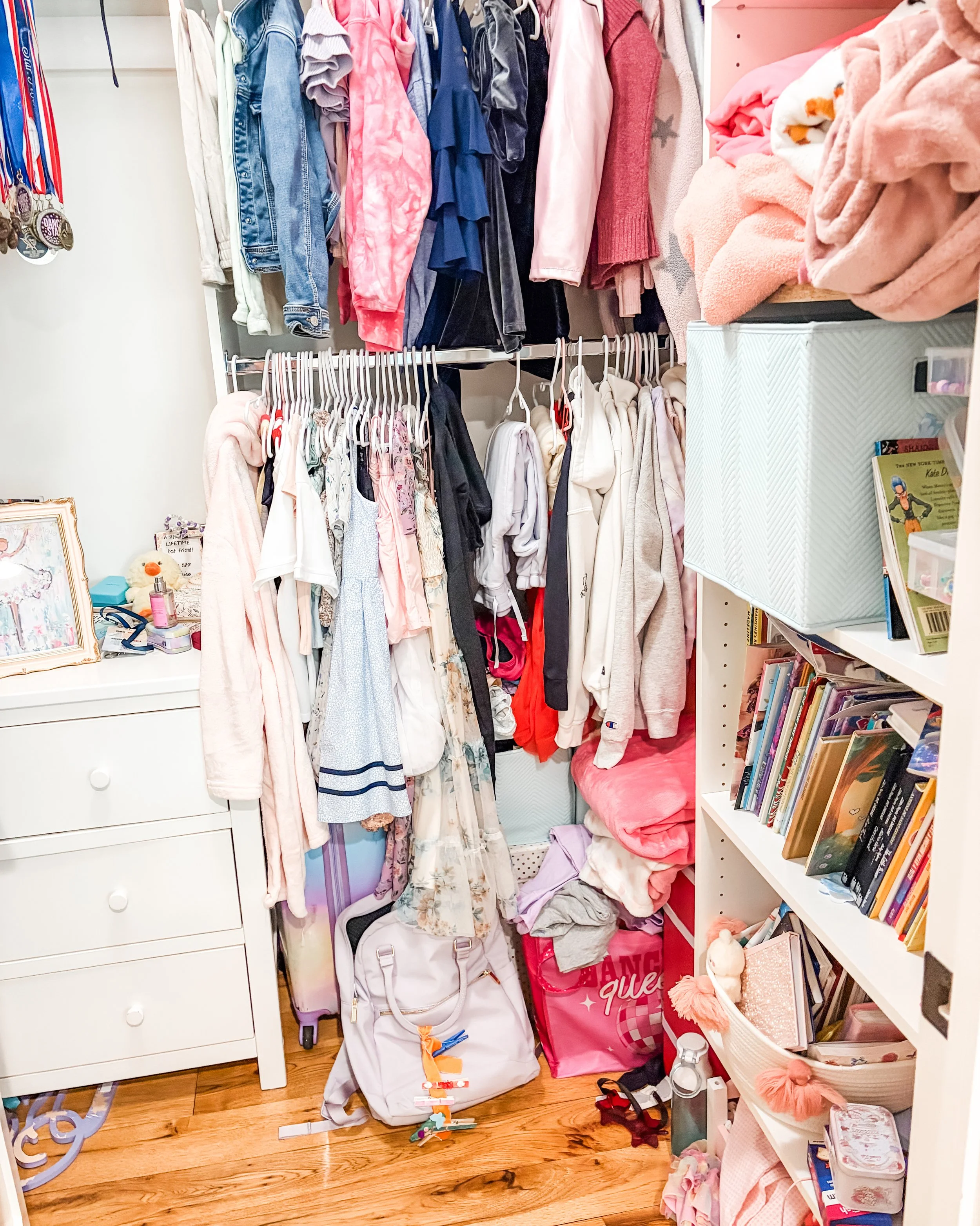 A closet with clothes hanging on a rack, shelves filled with folded clothes, books, and various personal items, including a white dresser with medals hanging from it, and a pink storage bin labeled 'dance' on a wood floor.