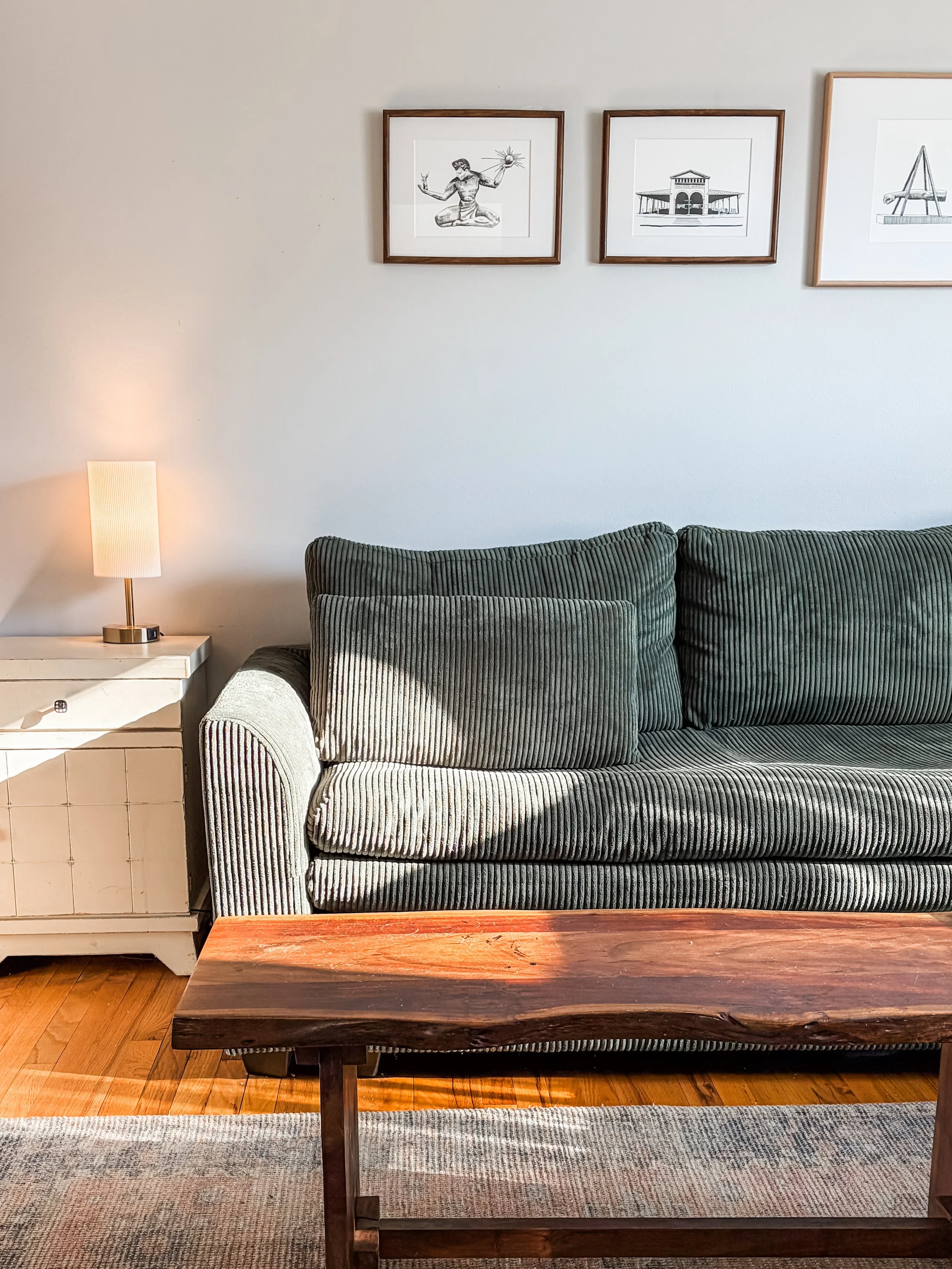 Living room scene featuring a green sofa, a white side table with a lamp, three framed art prints on the wall, a wooden coffee table, and a multicolored area rug.