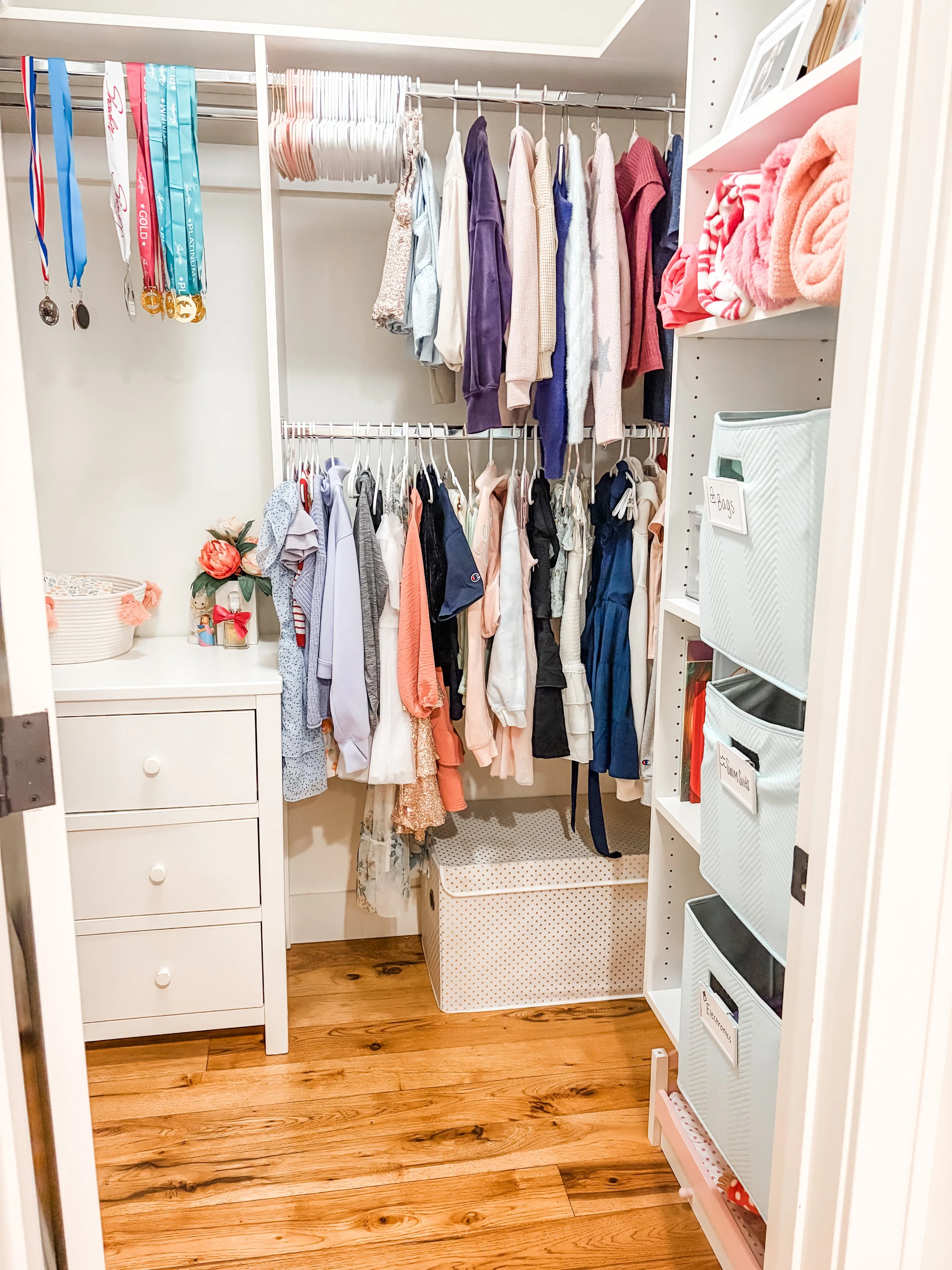 A walk-in closet with clothing hanging on rods, shelves with folded towels, and storage bins. The floor is wooden.