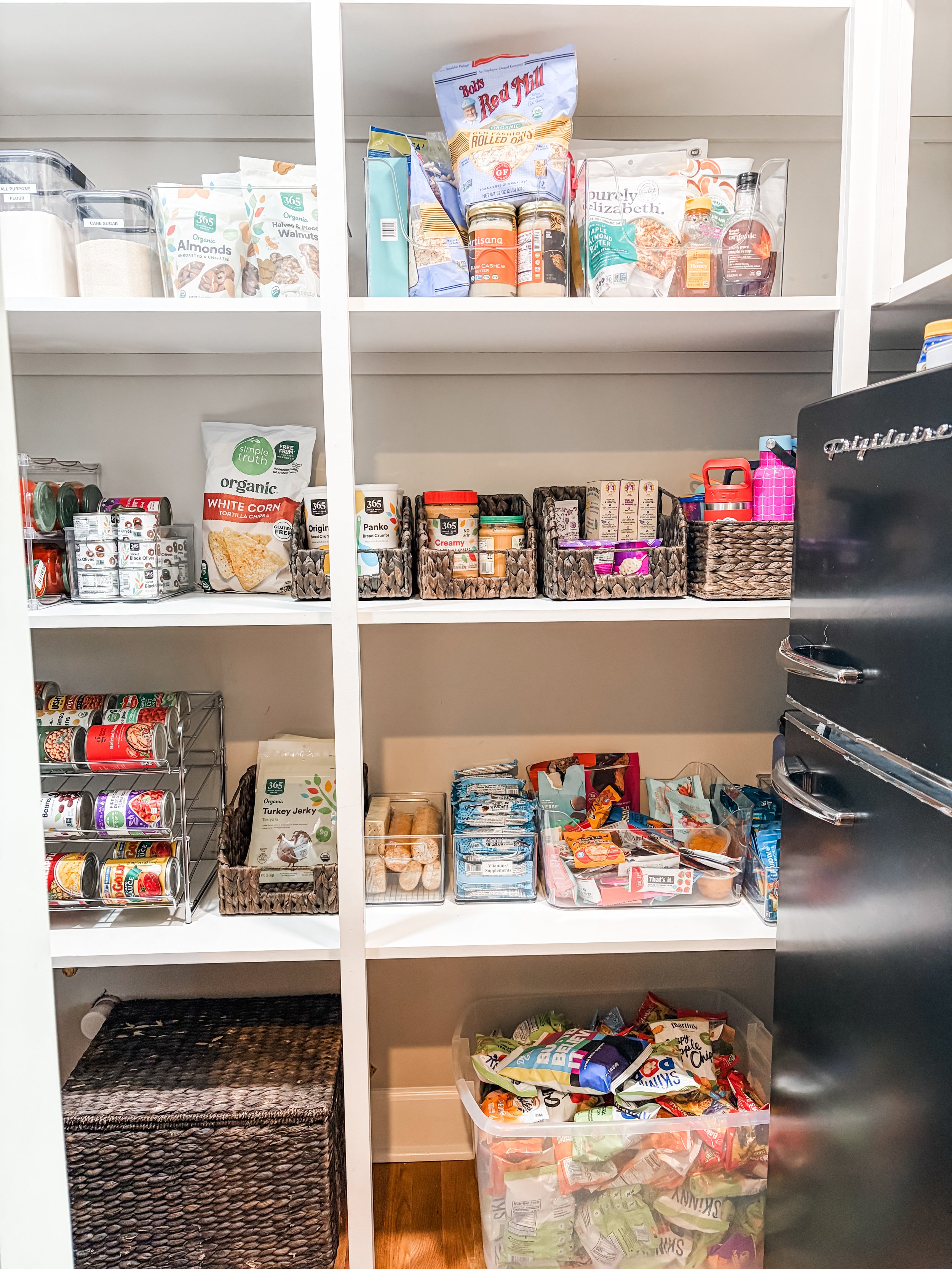 A pantry shelf with various food items, snacks, canned goods, and household supplies organized in baskets and containers.