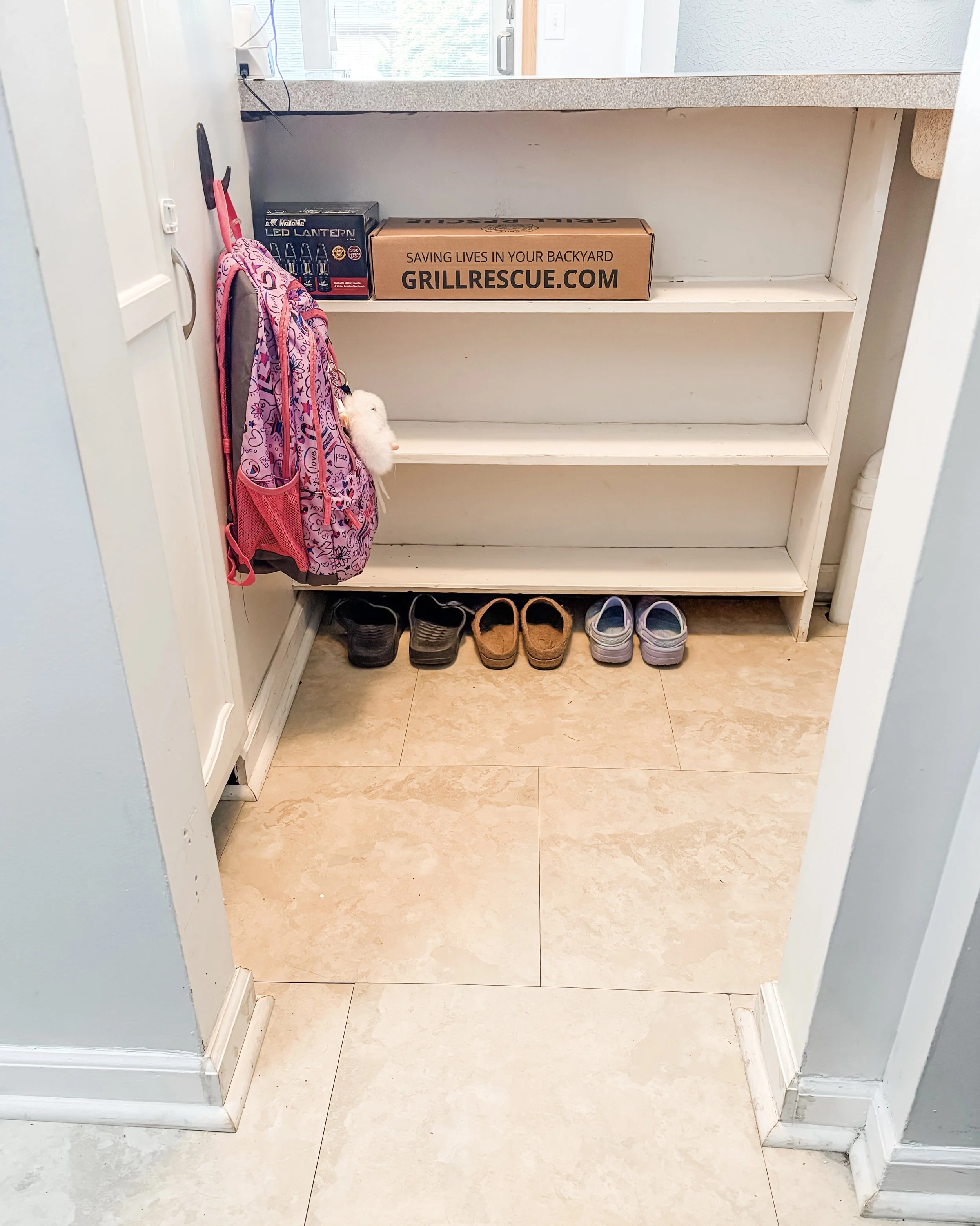 A view of a small hallway or entryway with beige tile flooring, white walls, and a white wooden shoe rack with three shelves. Three pairs of shoes are neatly arranged on the bottom shelf. The middle shelf is empty, and the top shelf holds a pink back