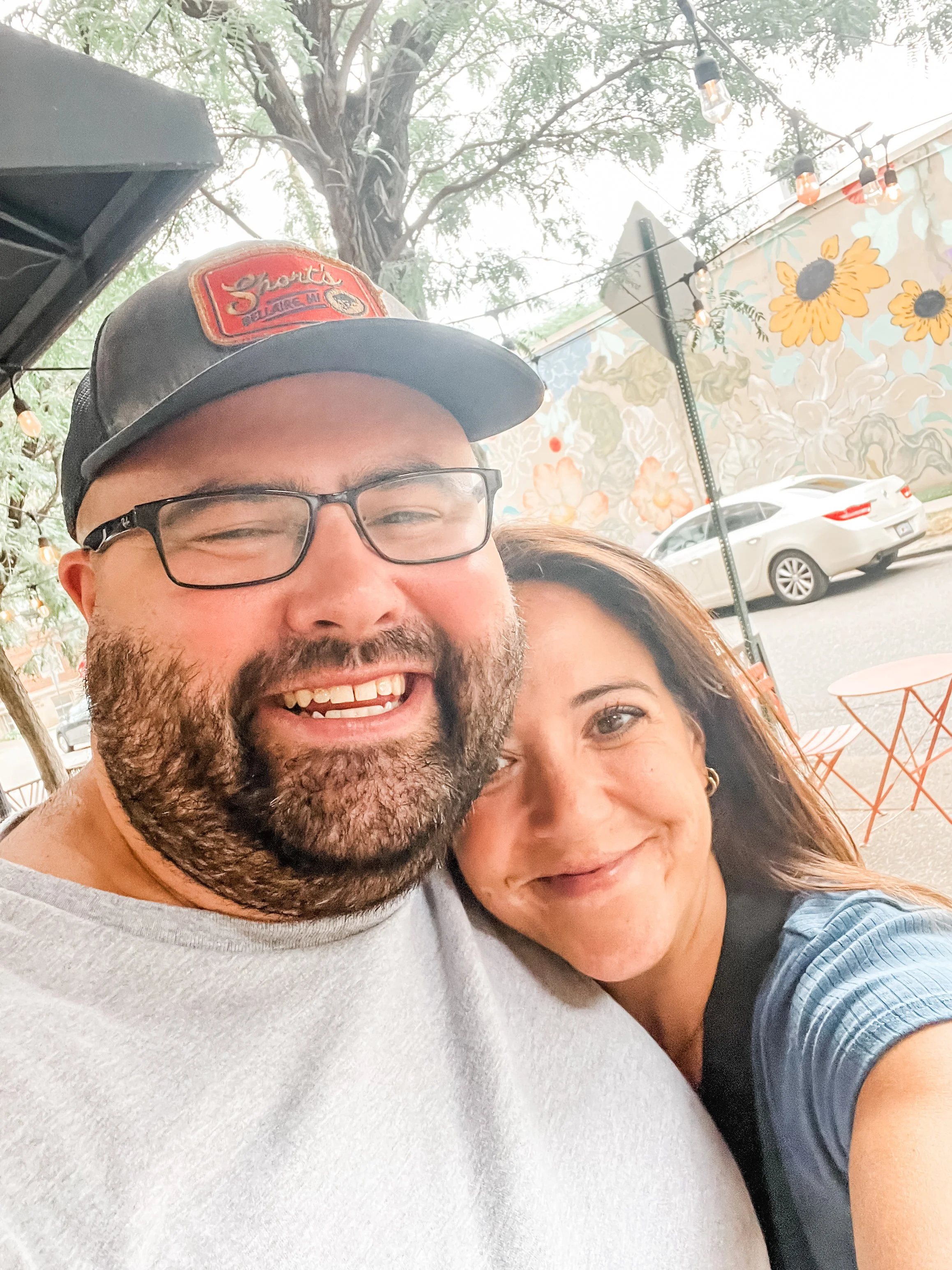 Close-up of a man and woman smiling outdoors, with string lights and a painted floral wall in the background.