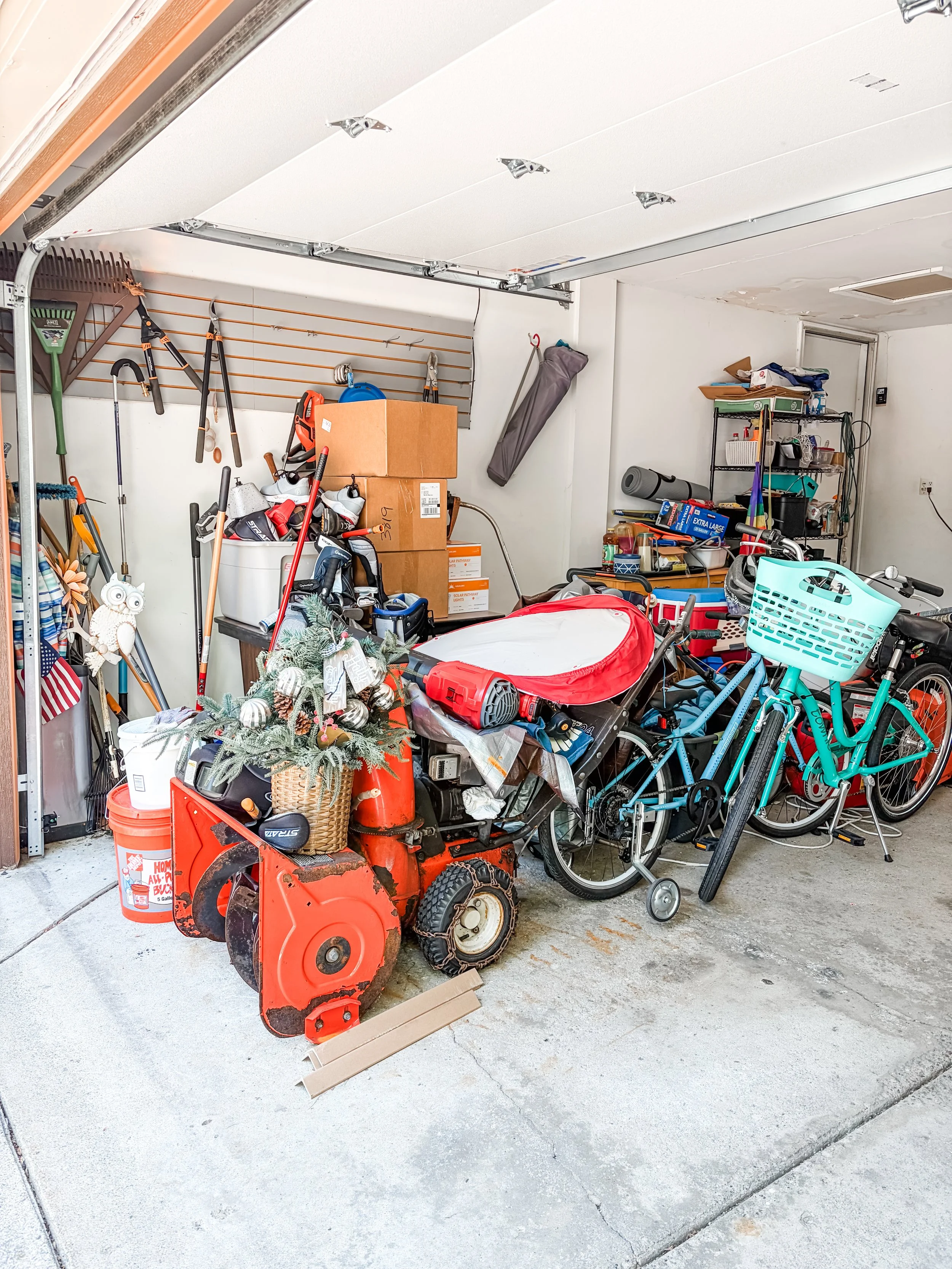 A cluttered garage with various tools, boxes, bicycles, and a red snow blower.