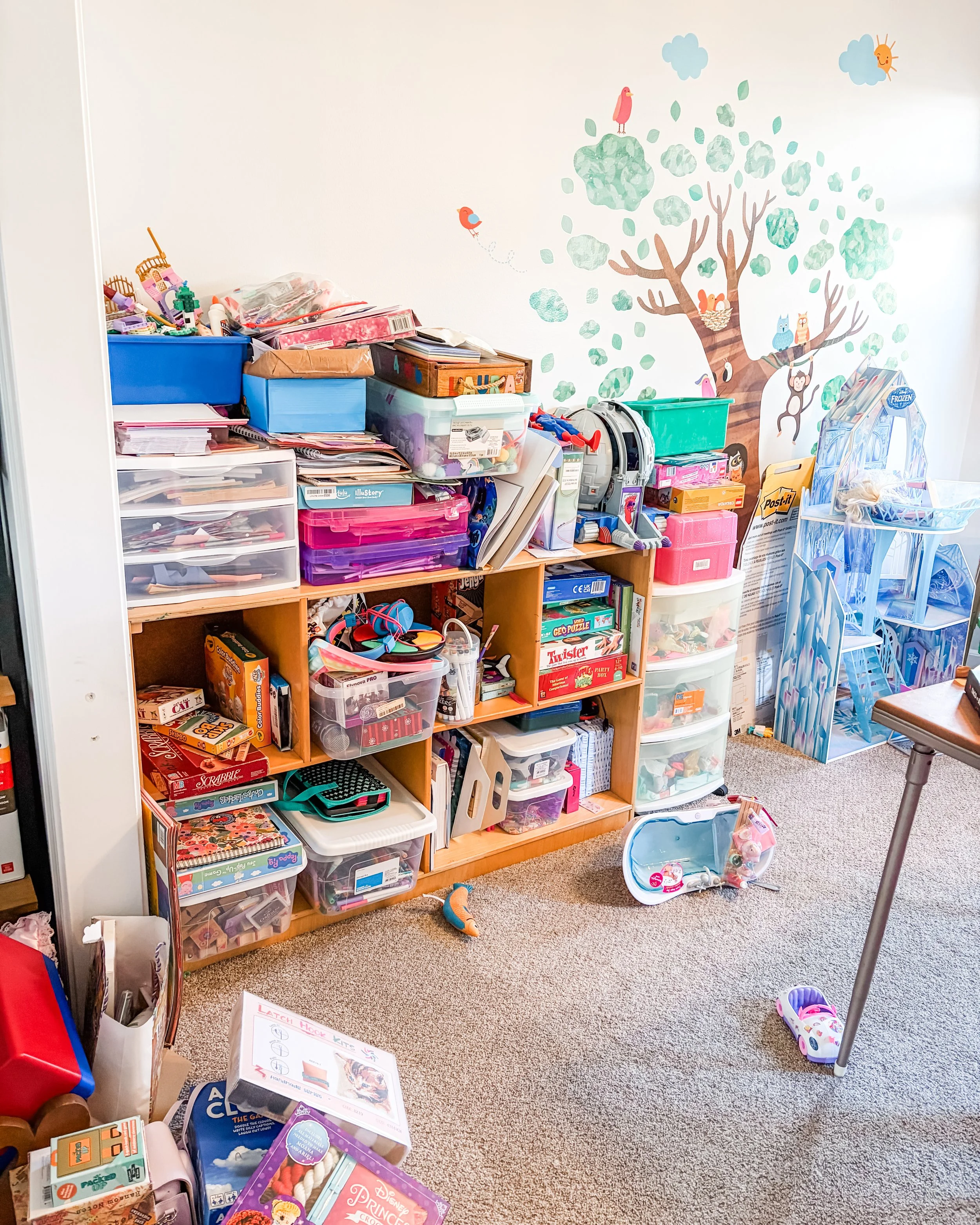 A cluttered room with shelves filled with books, board games, toys, and craft supplies. A large wall decal features trees, clouds, birds, a butterfly, an owl, and a monkey. The room has beige carpet and a stool with a fabric seat facing away from the