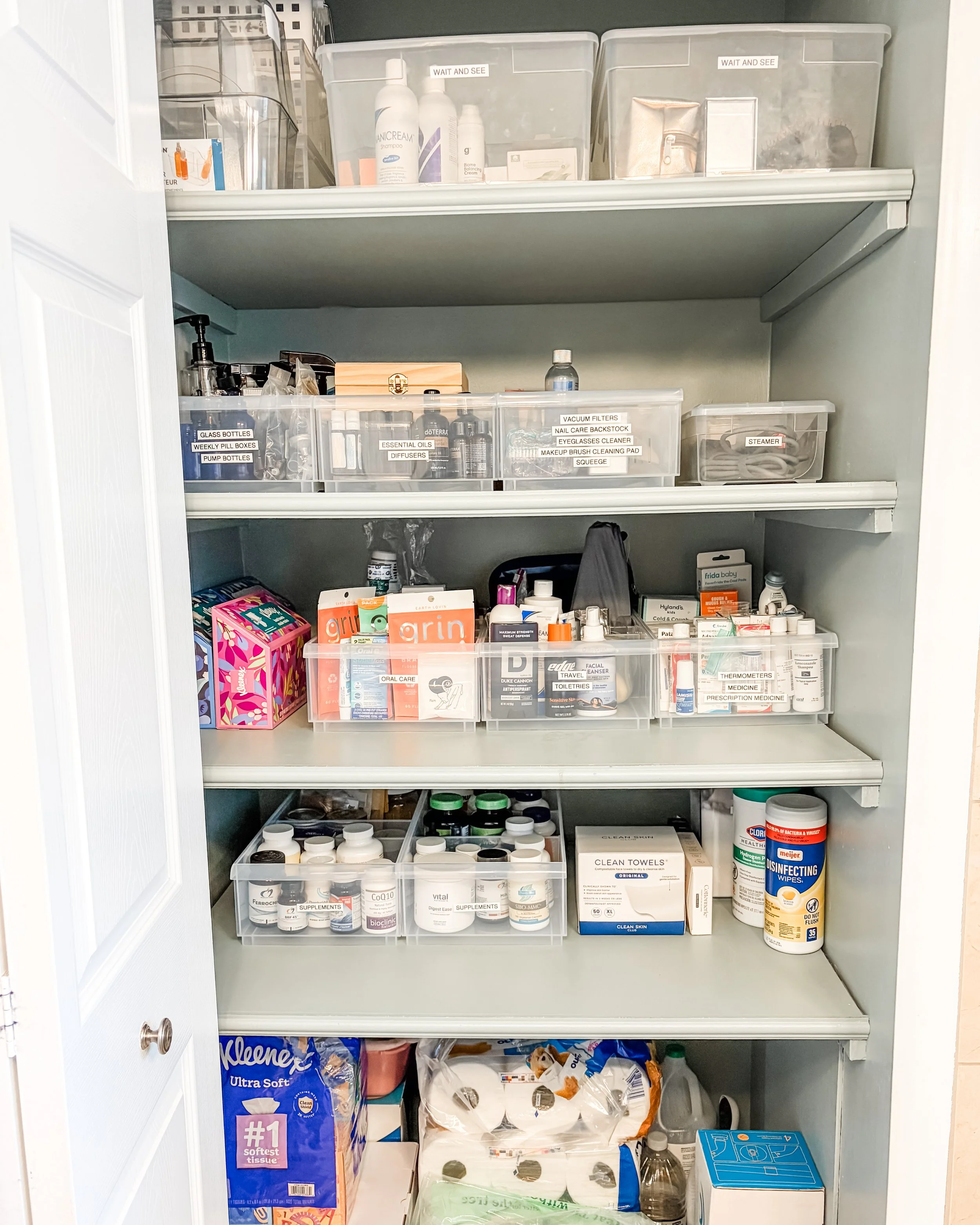 A closet with three shelves filled with various household supplies including bottles, containers, tissues, disinfecting wipes, and medical supplies, organized in clear labeled bins and boxes.