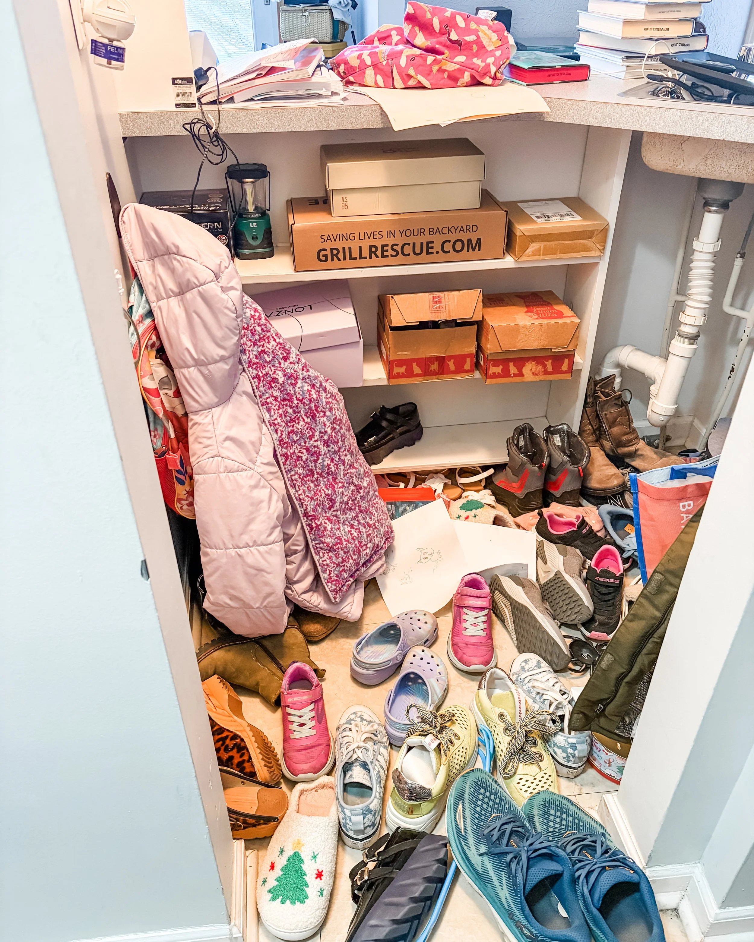 A cluttered entryway or small closet area with various shoes on the floor, some scattered and others organized. Shoes include sneakers, boots, and sandals. There are boxes, clothing, and miscellaneous items on shelves and the floor, including a pink 