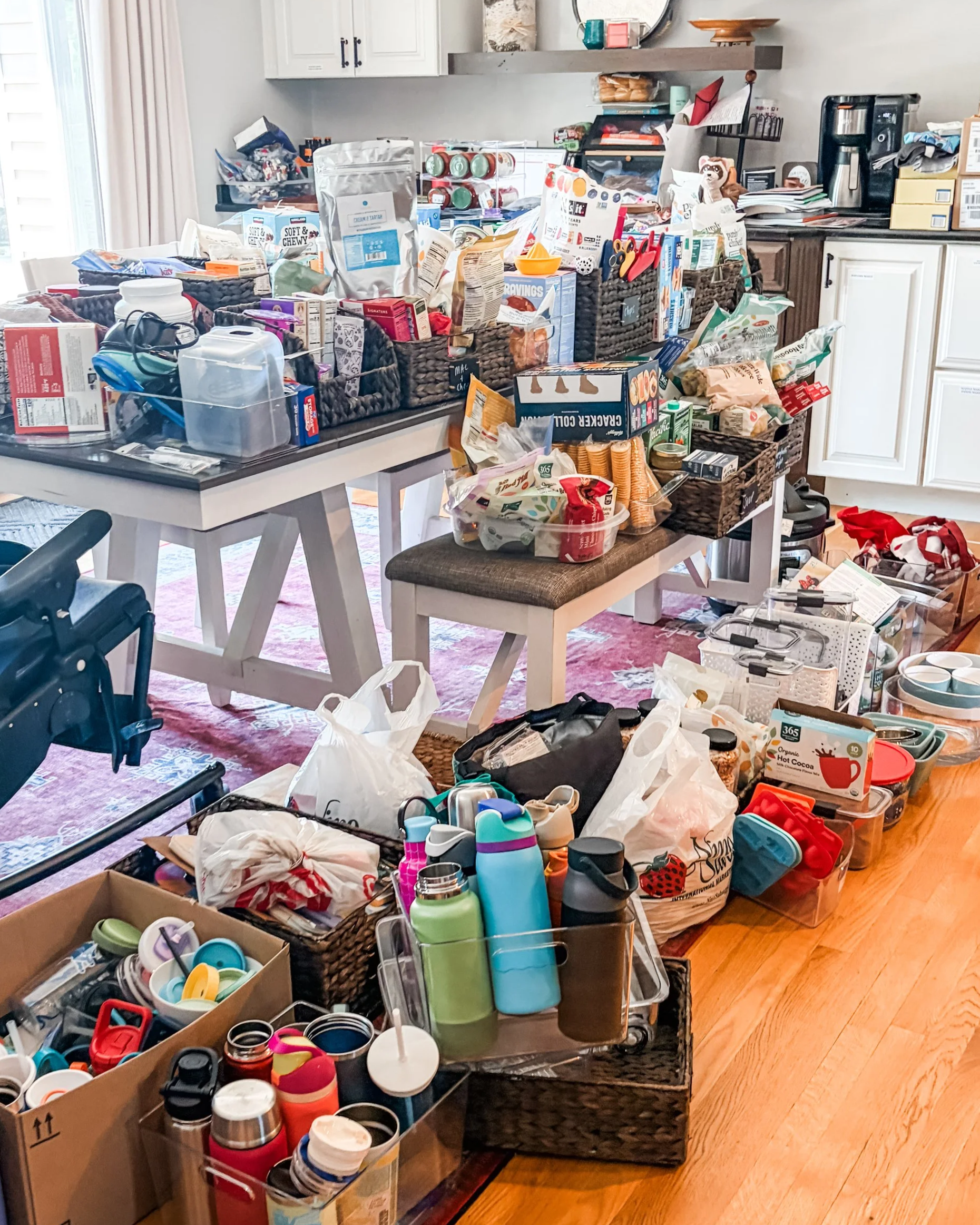 A cluttered kitchen with various items on a table, baskets, boxes, and shelves filled with snacks, kitchenware, and miscellaneous household items.
