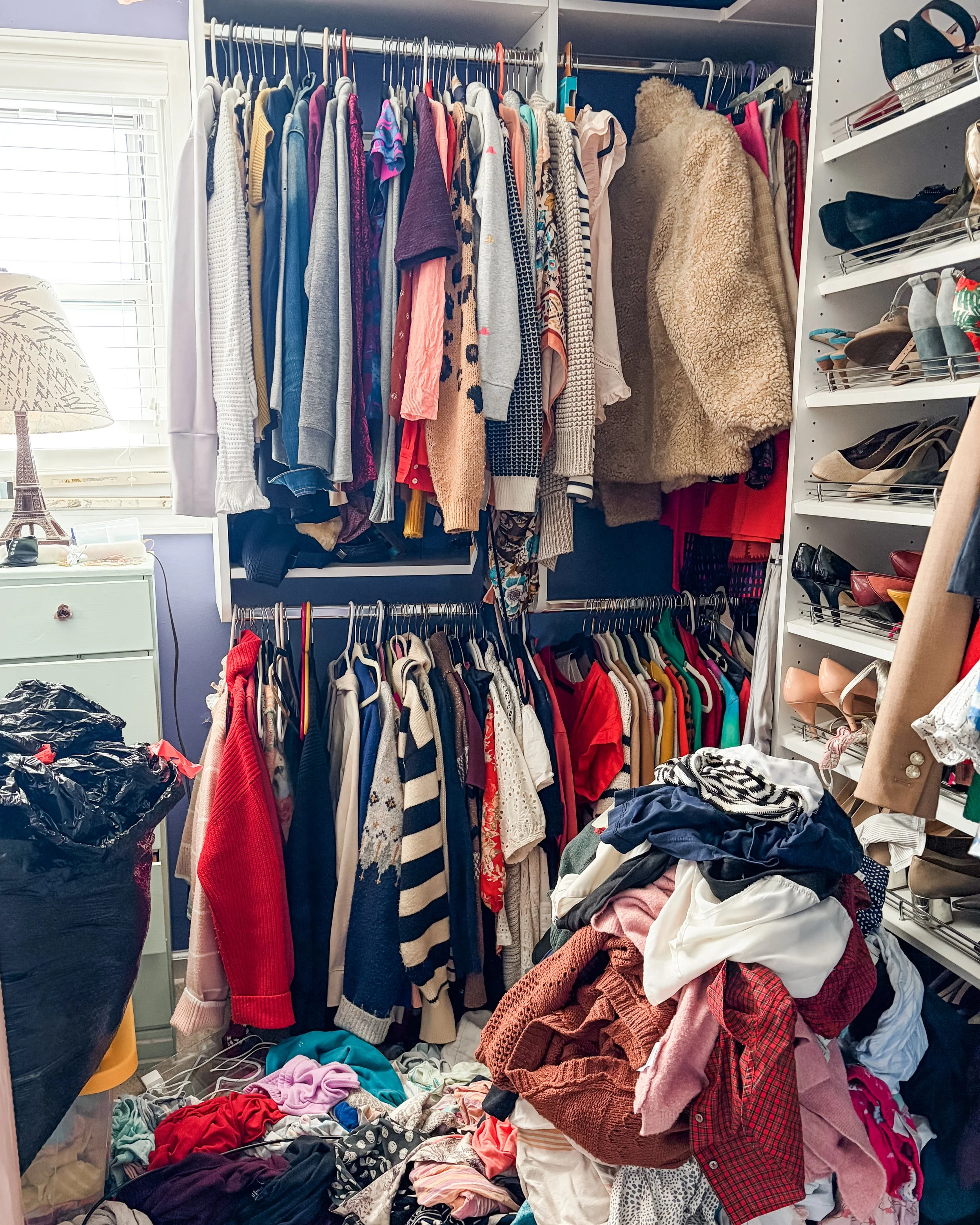 Clothes closet filled with hanging and piled clothes, including sweaters, shirts, and dresses, with a pile of clothes on the floor and scattered on furniture.