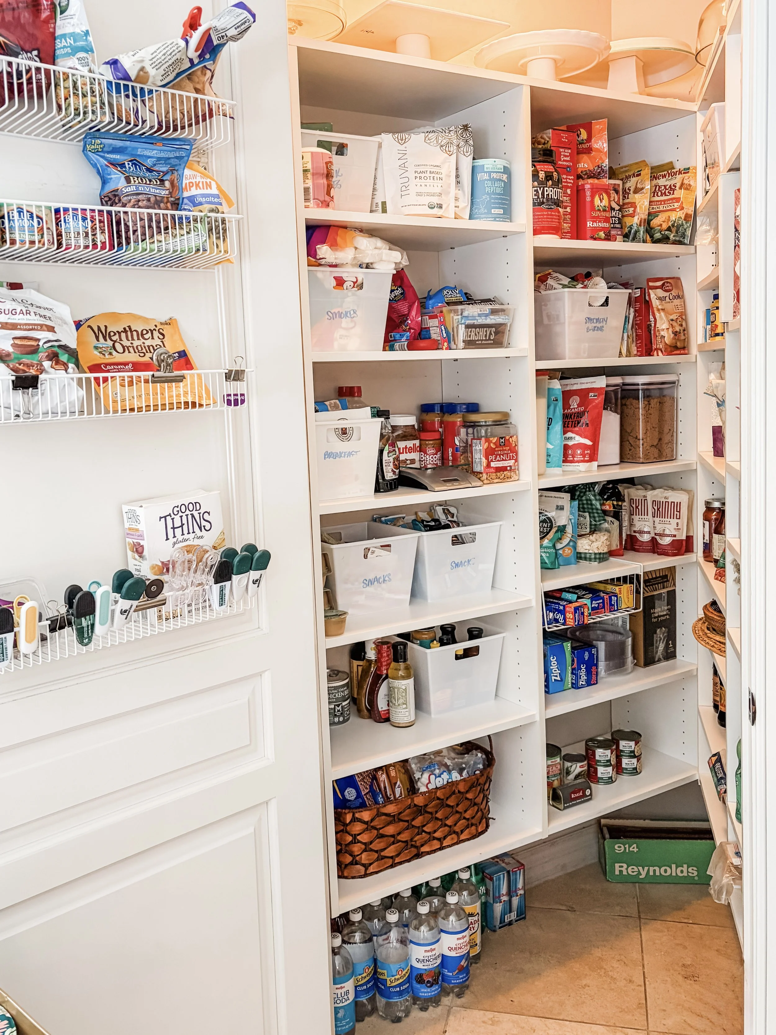 A pantry with white shelving filled with snacks, canned goods, bottled drinks, and baking supplies. The left side features a closet with wire baskets containing packaged snacks and a brochure. The right side shelves hold various food items, jars, bot