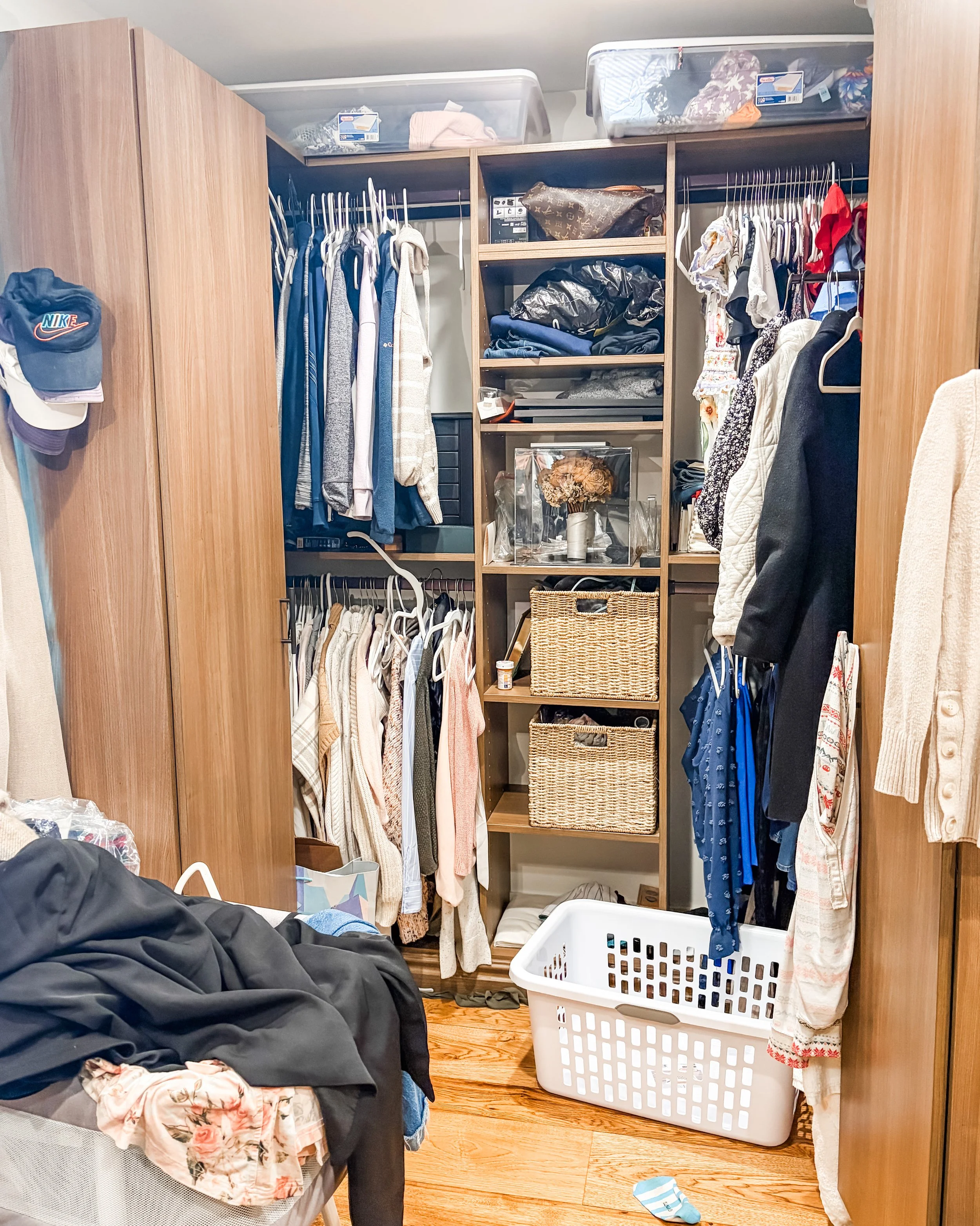 Cluttered walk-in closet with clothes hanging on rods, clothing storage bins on top shelves, and a large white laundry basket on wood floor, with various clothing items and a black jacket on bed.