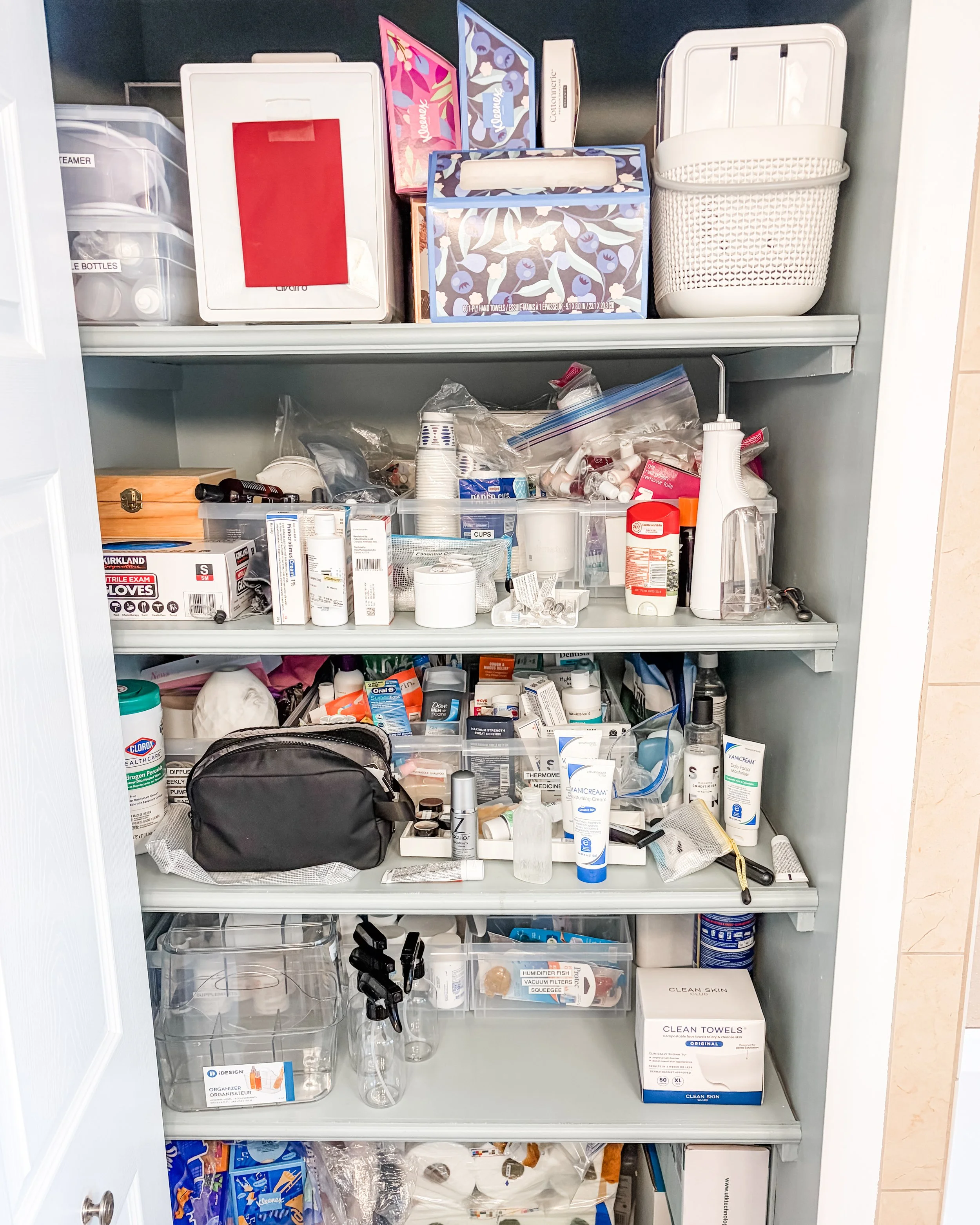 A closet shelf with various personal care and household items including medications, skincare products, a black toiletry bag, ziplock bags filled with small items, a box of clean towels, and miscellaneous containers.