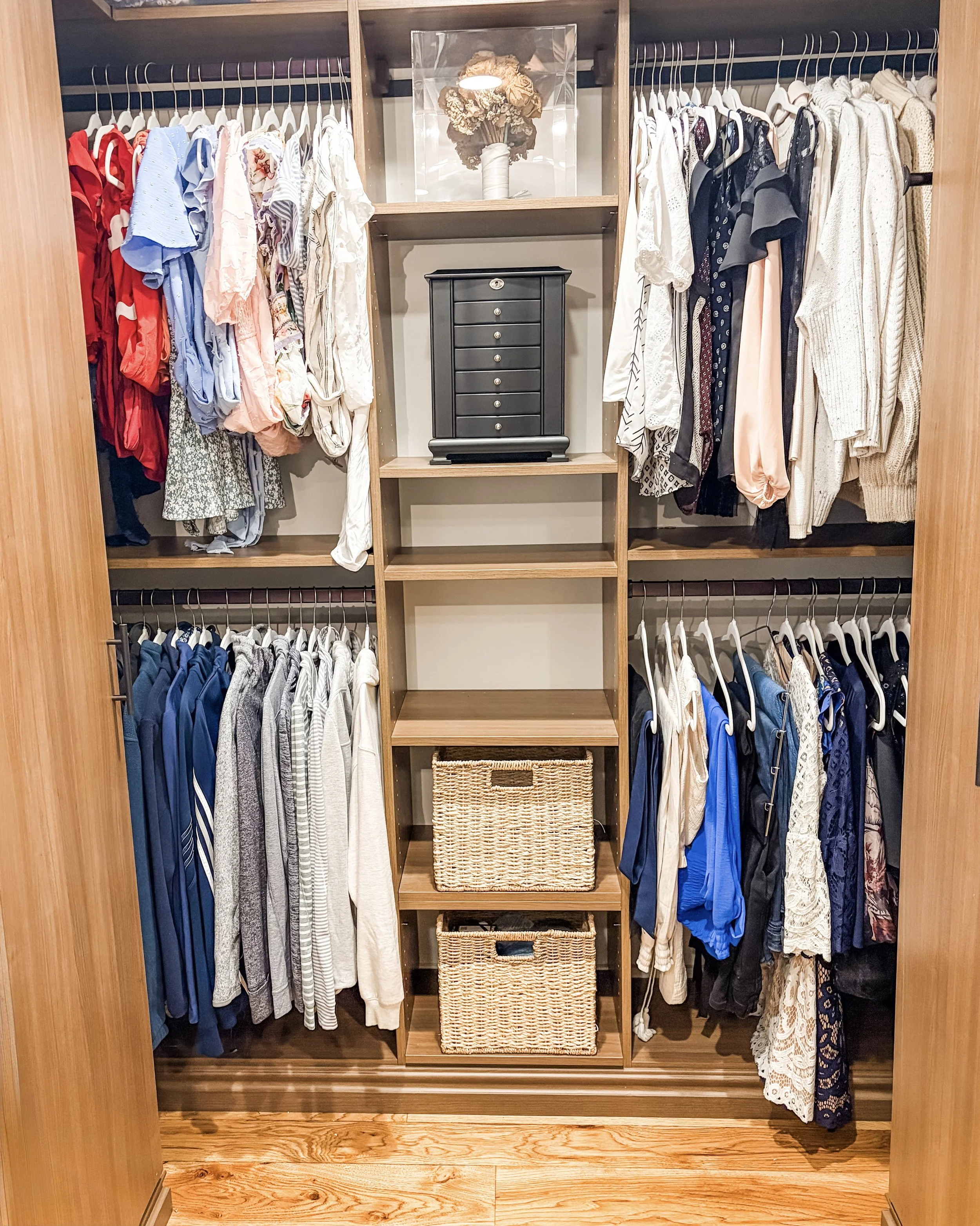 A neatly organized walk-in closet with hanging clothes on both sides, including dresses, shirts, and pants. There are two woven baskets on the lower shelves and a small black jewelry or storage box in the center. A decorative floral arrangement in a 