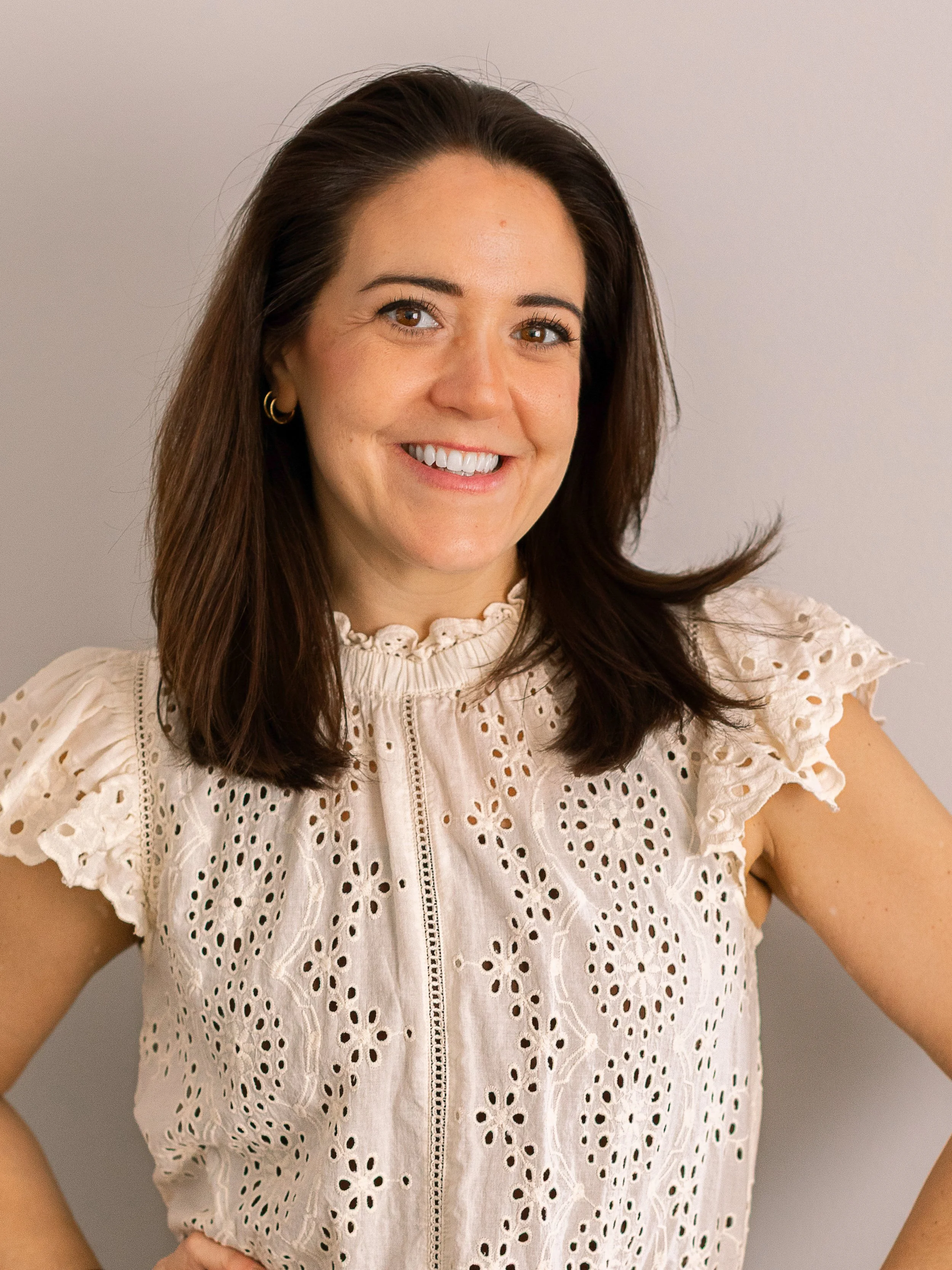 A woman with brown hair smiling, wearing a cream-colored eyelet blouse with ruffled sleeves and a high neckline, standing against a plain light-colored background.