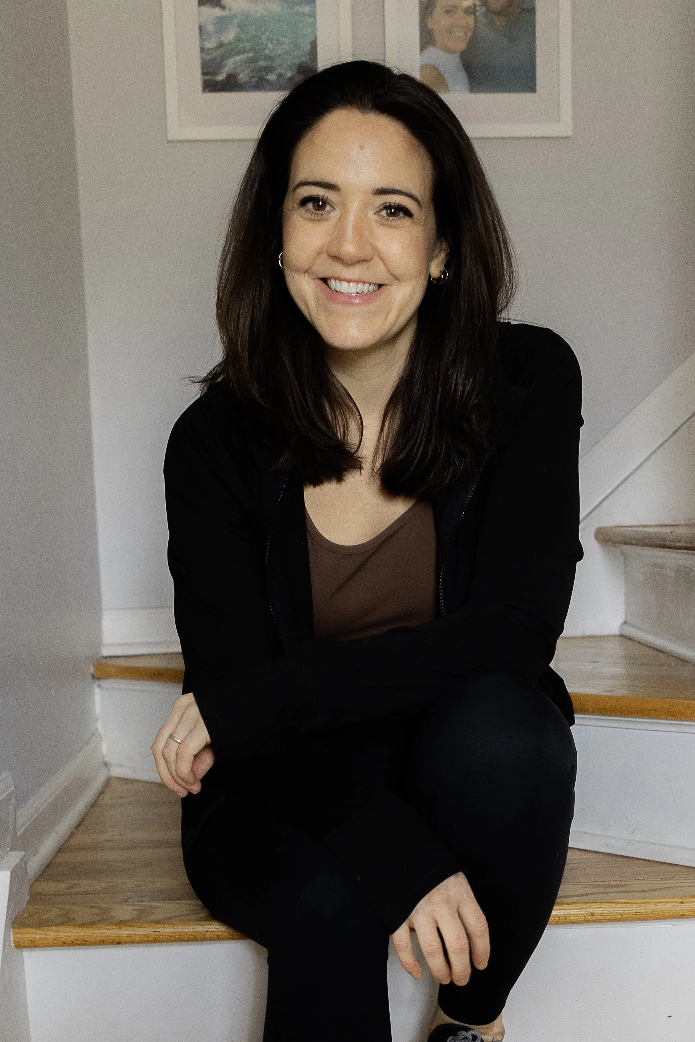 A woman with dark brown hair smiling and sitting on wooden stairs in a house, with framed photographs of people on the wall behind her.