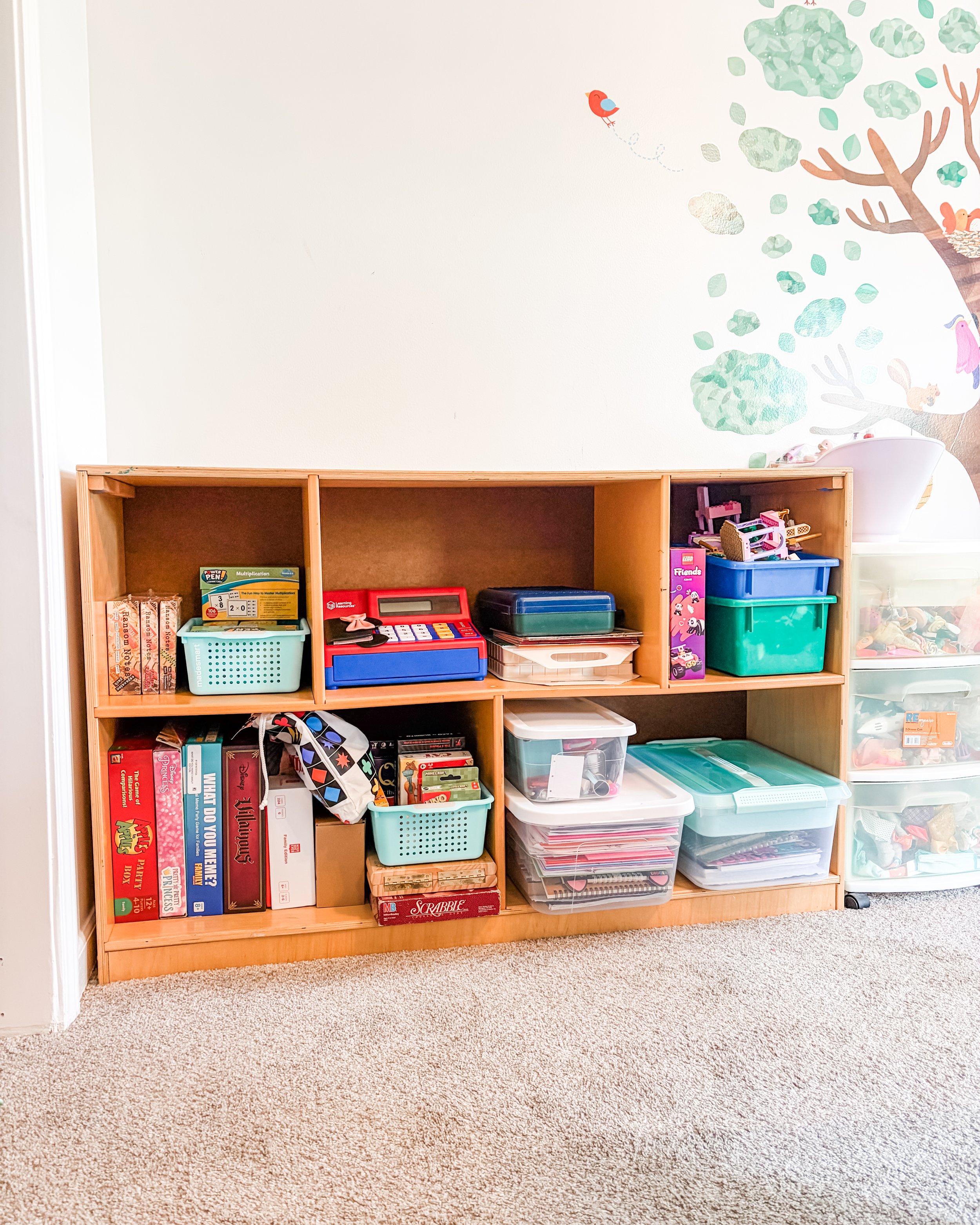 Wooden bookshelf with toys, books, and storage bins against a wall with a tree and bird mural