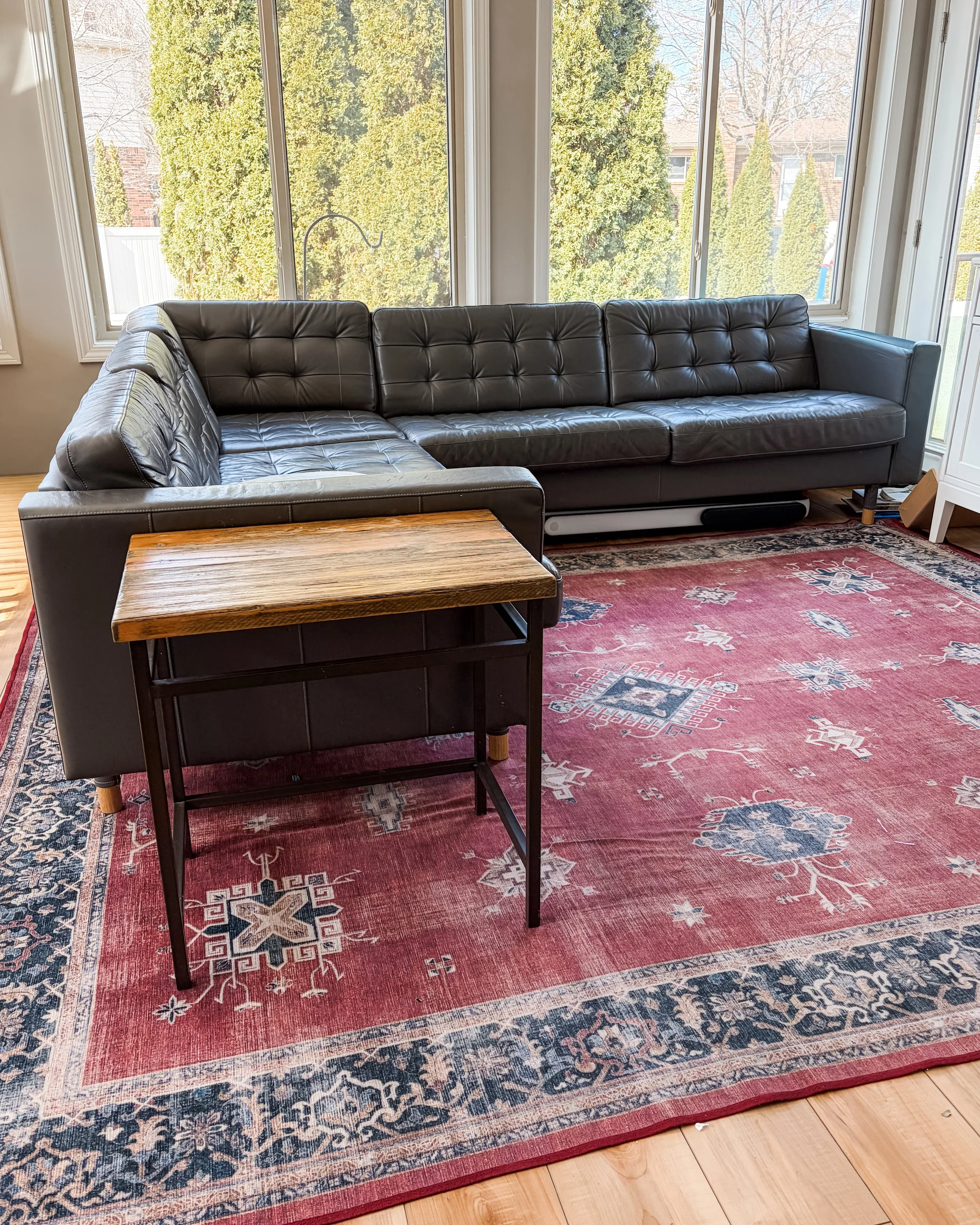 A living room with a large L-shaped black leather couch placed on a red and blue patterned area rug in front of three large windows showing trees and residential buildings outside. There is a wooden coffee table in front of the couch and hardwood flo