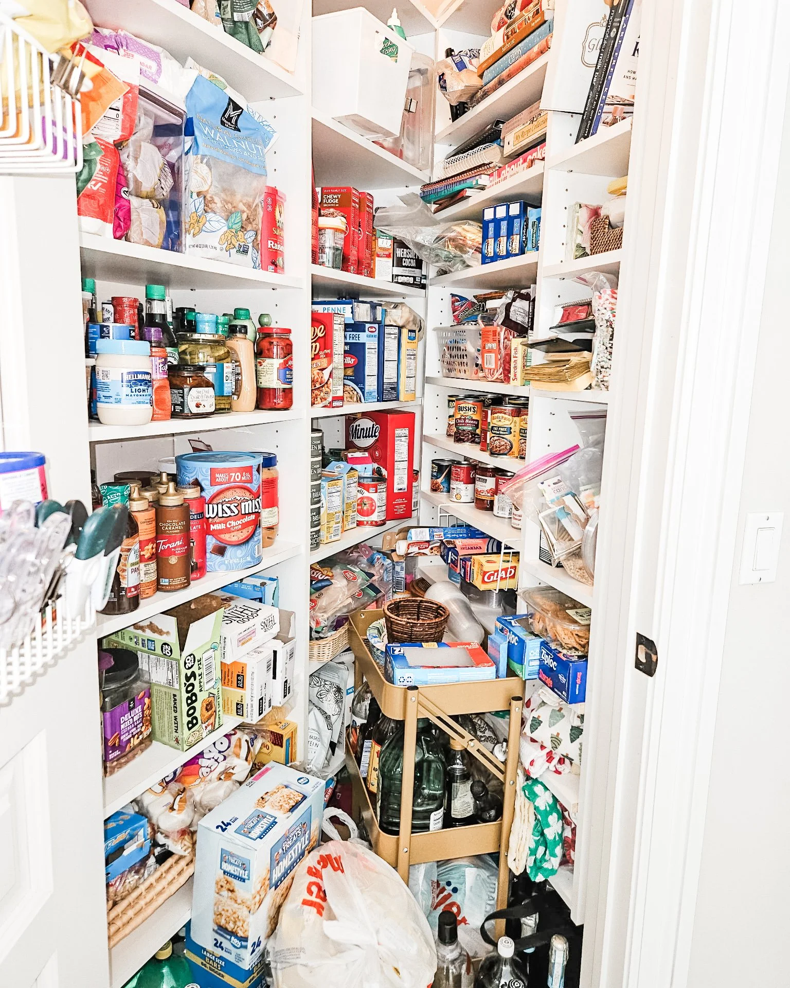 A small storage pantry filled with food items, including canned goods, boxes of cereal, bottles, and miscellaneous snacks and pantry essentials on white shelves.