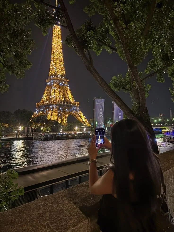 A woman taking a photo of the Eiffel Tower at night, illuminated with lights, across the river. There are trees and city lights nearby.