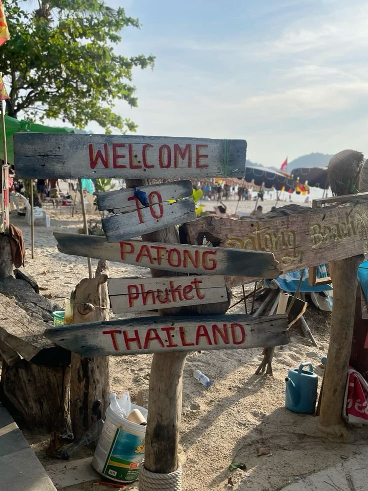 A weathered wooden sign on the beach welcoming visitors to Patong Beach in Phuket, Thailand. The sign is handmade, with red and black lettering, and is partially surrounded by beach supplies and natural debris. In the background, there are people, umbrellas, and the ocean under a partly cloudy sky.