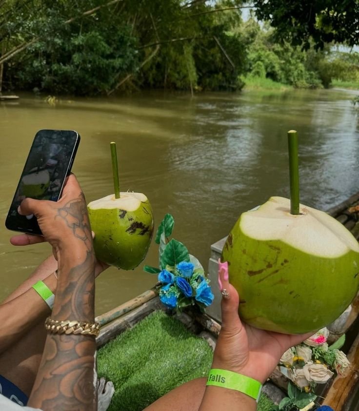 Two people holding large green coconuts with straws near a river, with one person taking a photo with a smartphone. There are blue flowers decorated on the railing or nearby area.