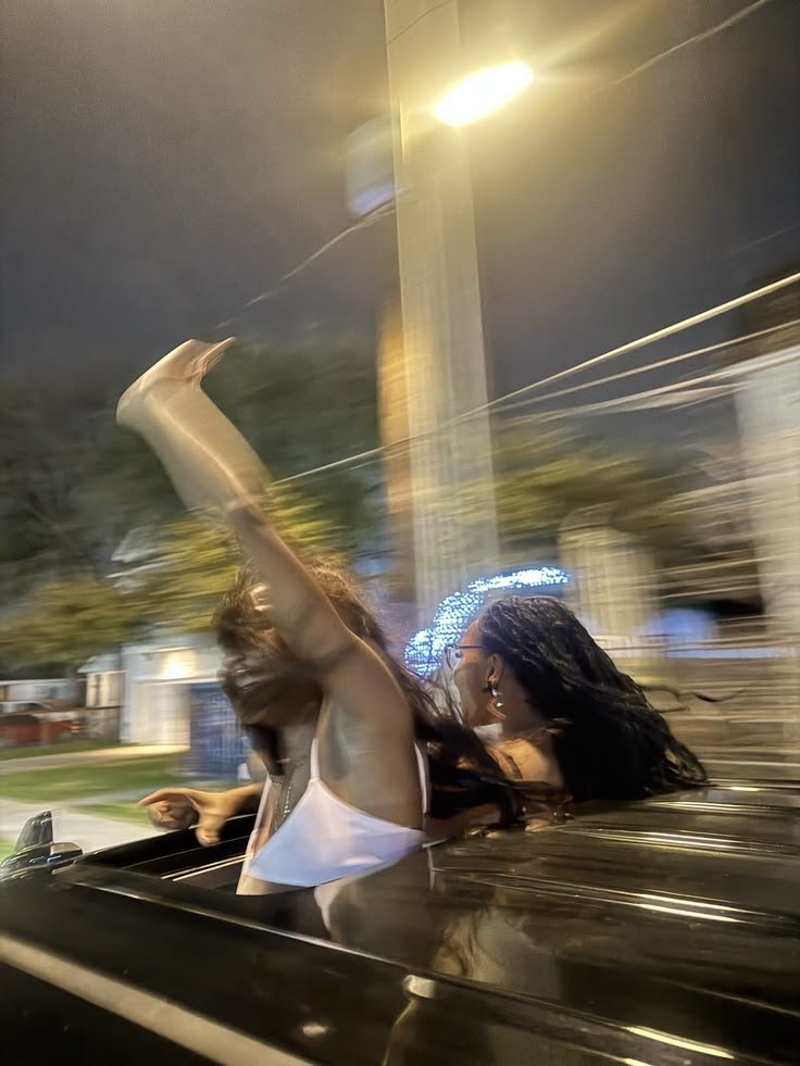 Two women are riding in a moving car at night, with one of them raising her arm. The background shows blurred streetlights and houses, indicating motion.