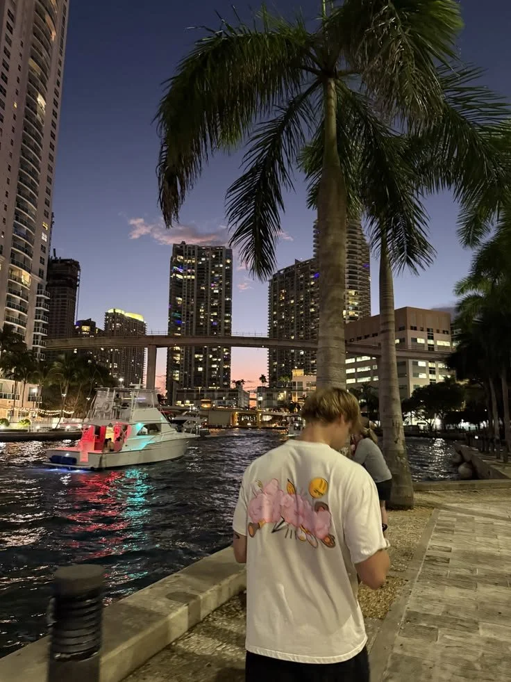 A person wearing a white T-shirt with pink flamingos and a yellow emoji on their back, walking along a waterfront promenade at sunset. Tall buildings, palm trees, and a boat on the water are visible in the background.