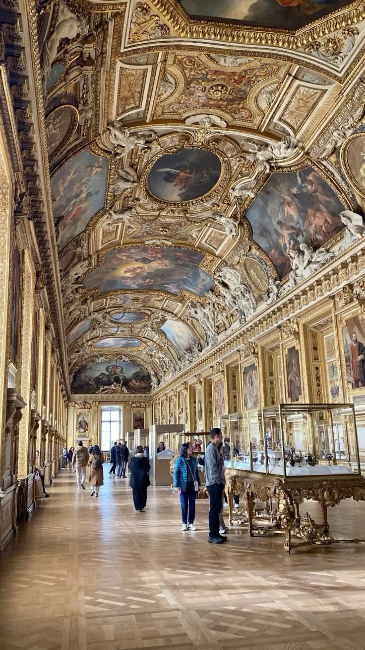 A grand, ornately decorated museum hall with large ceiling murals, framed paintings along the walls, and glass display cases showcasing artifacts. Visitors are walking and viewing exhibits.