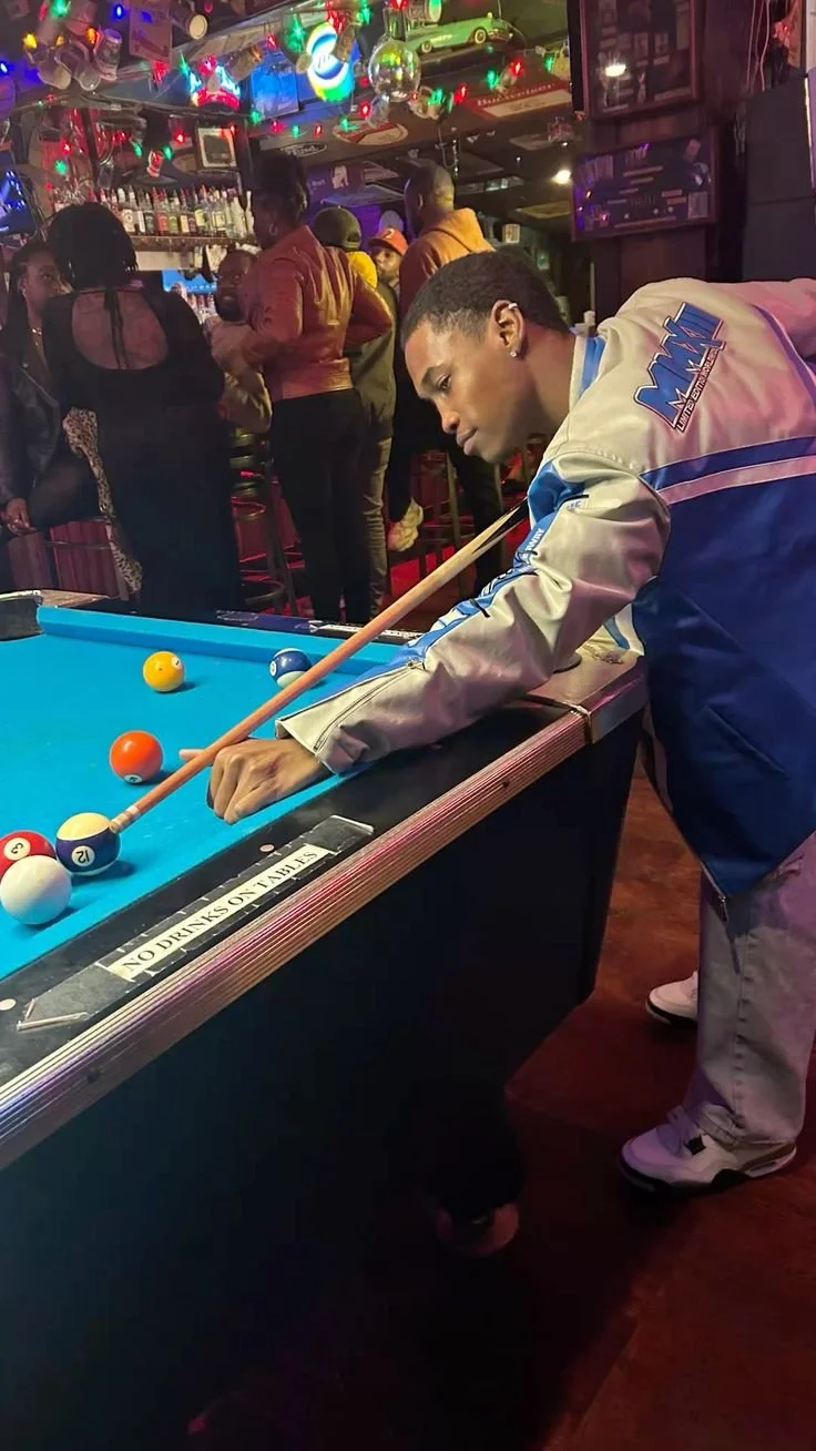 A young man playing pool at a bar, with multiple people socializing in the background, festive lights, and a disco ball overhead.