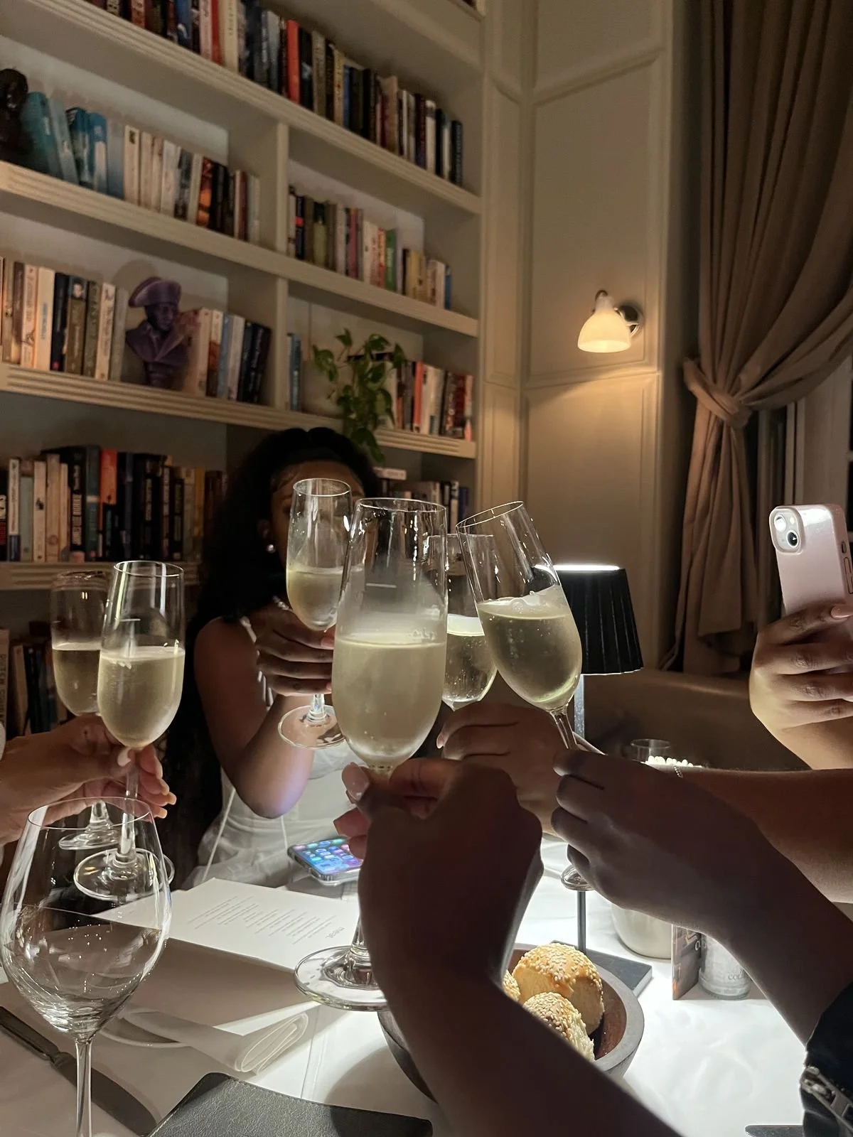 People raising glasses of champagne in a toast at a dinner party in a cozy, well-lit room with bookshelves and curtains.