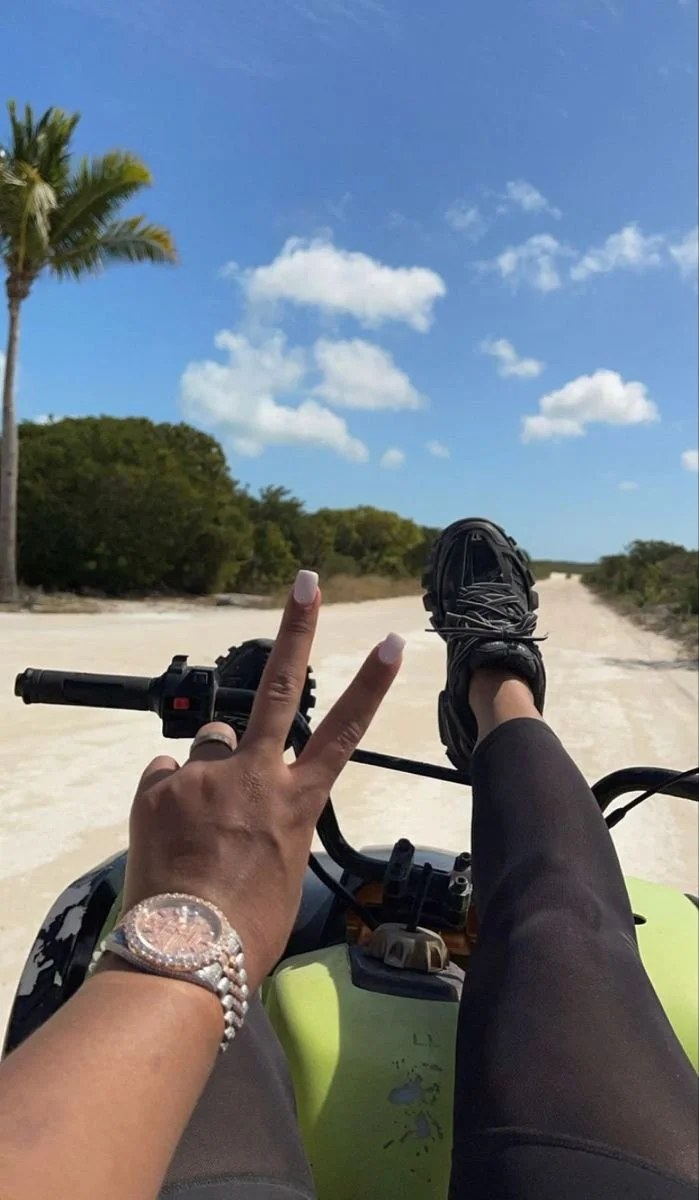 Person on a green dirt bike taking a selfie while relaxing on the handlebars, wearing black athletic pants and a watch, on a sandy trail with trees and a blue sky with clouds.