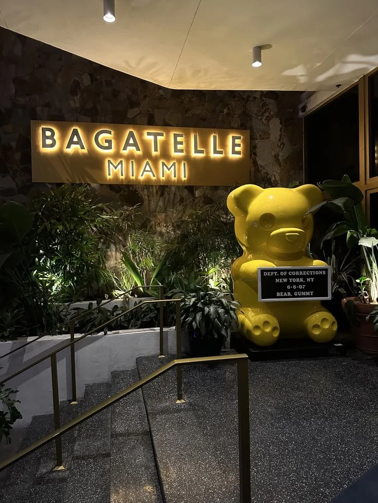 Indoor scene of the entrance or lobby with a large illuminated sign saying "Bagatelle Miami" and a yellow bear-shaped GUMMY toy with a label from the Department of Corrections in New York, NY, identifying it as a bear gummy, surrounded by plants and greenery.