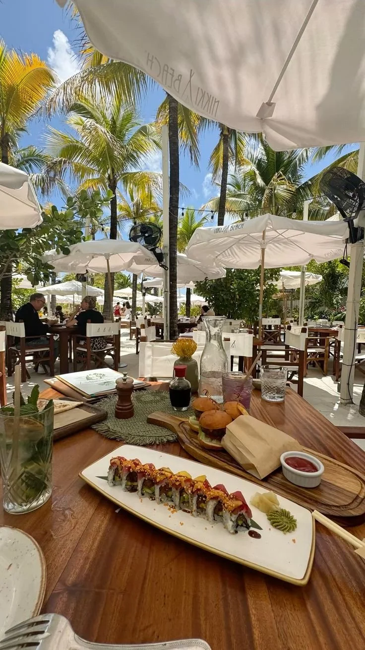 A table at a beachside restaurant with sushi, drinks, and condiments under white umbrellas, surrounded by palm trees and a partly cloudy sky.