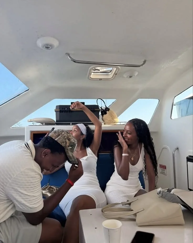 Three friends enjoying time together on a boat, sitting on a blue bench. Two women in white dresses are smiling and one man in a white shirt and Camouflage cap appears to be laughing or feeling emotional, with his hand on his forehead. There are bags, a cup, and a smartphone on the table in front of them.