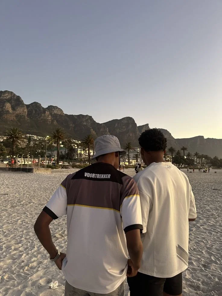 Two young men are standing on a sandy beach during sunset, with mountains and palm trees in the background, engaged in conversation.
