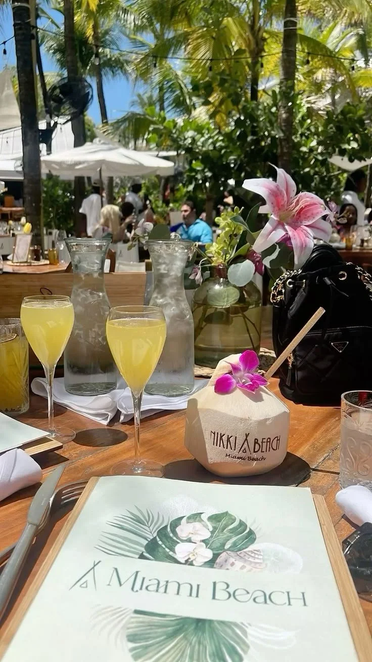 A restaurant table set outdoors at Nikki Beach in Miami Beach with flowers, two pitchers of water, three glasses of yellow drinks, a coconut with a straw and pink flower, a black bag, and a menu with tropical leaf design.