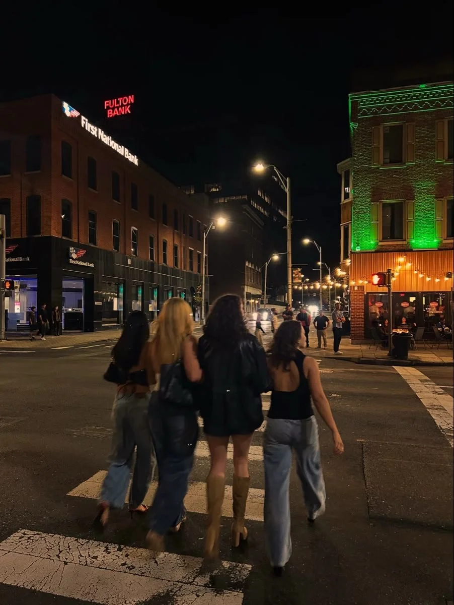 Four women crossing a city street at night, illuminated buildings and streetlights, with people at outdoor tables.