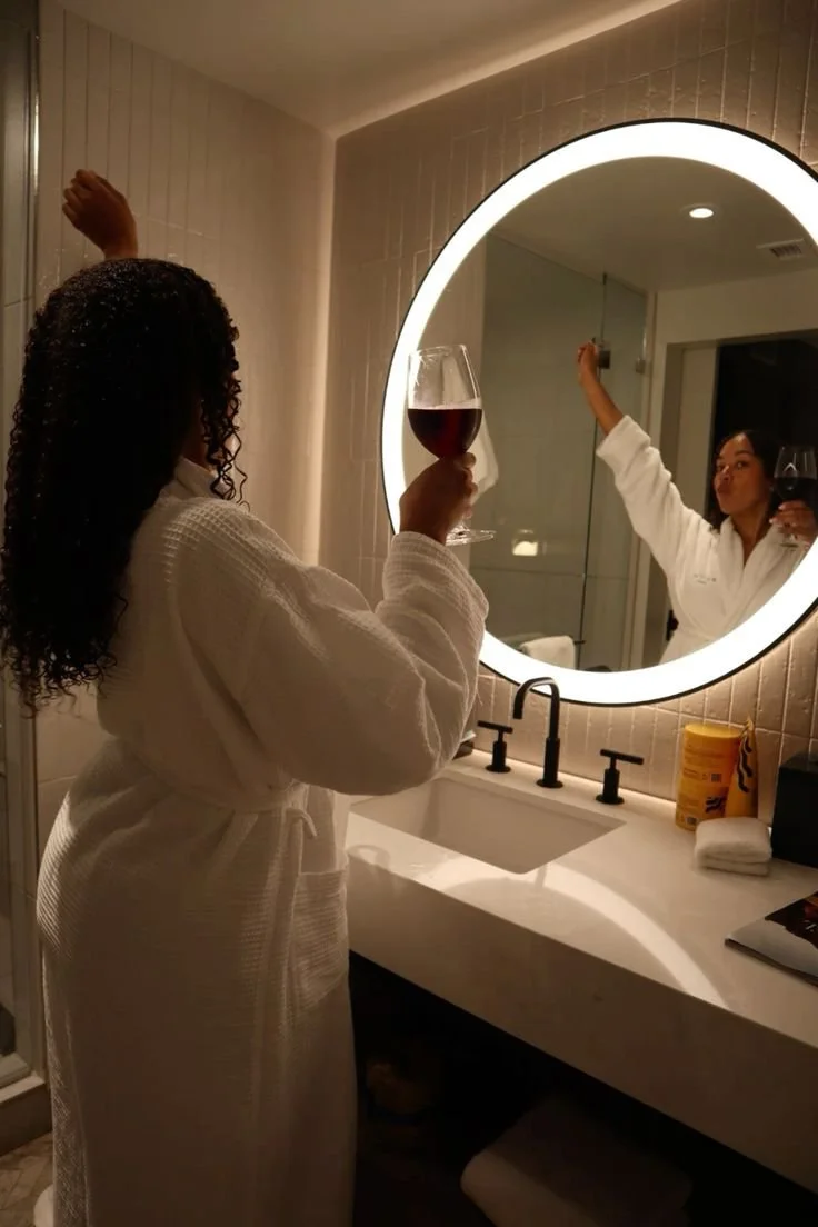 A woman in a white bathrobe holding a glass of red wine while looking at her reflection in a circular bathroom mirror, also holding a glass of red wine.