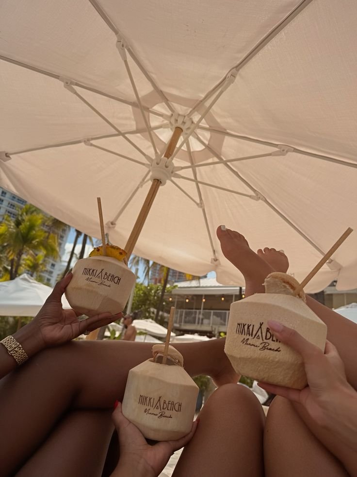 Three coconut bowls with straws, held by three people, under a large white beach umbrella at Nikki Beach Miami Beach.