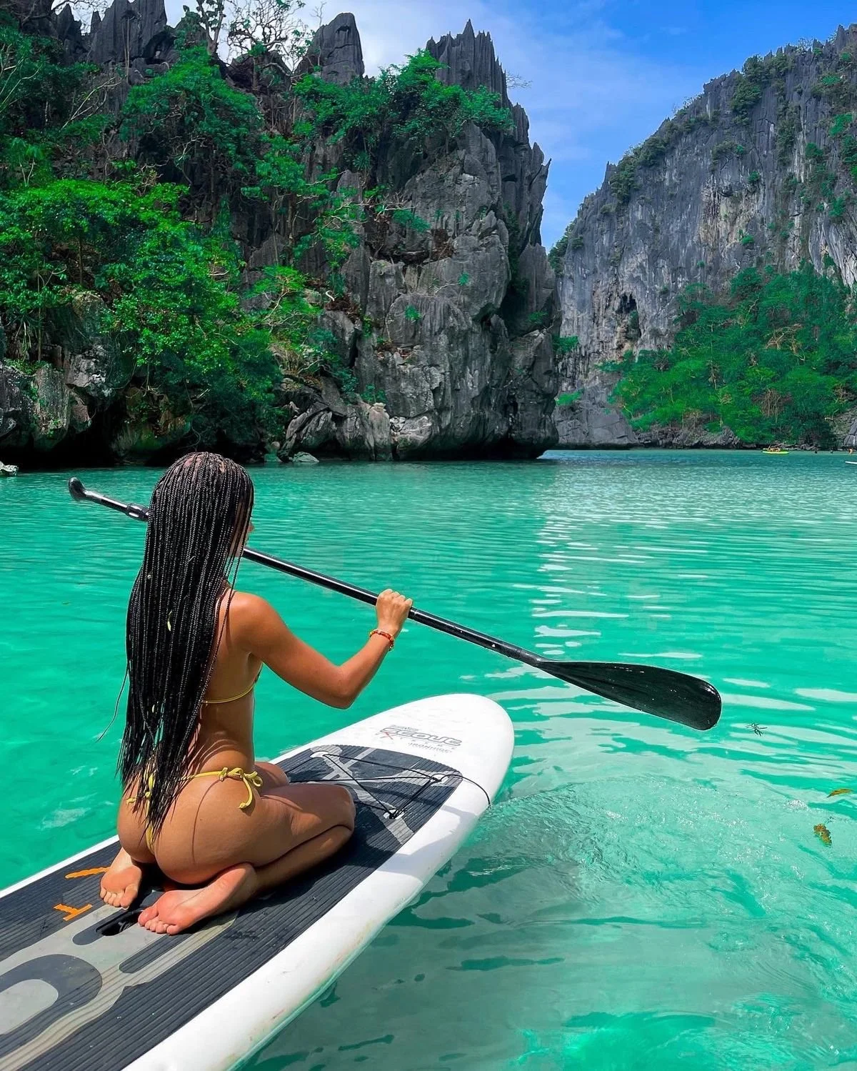 A woman with long braids kneeling on a paddleboard in turquoise water, holding a paddle, with lush green trees and high rocky cliffs in the background.