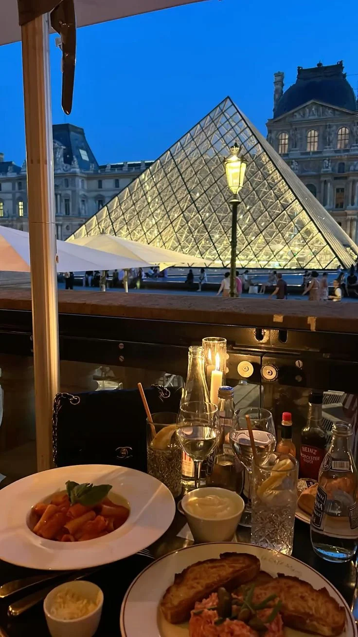 Indoor dining at a restaurant with a view of the Louvre Pyramid in Paris during evening; table set with pasta, garlic bread, drinks, and candles.