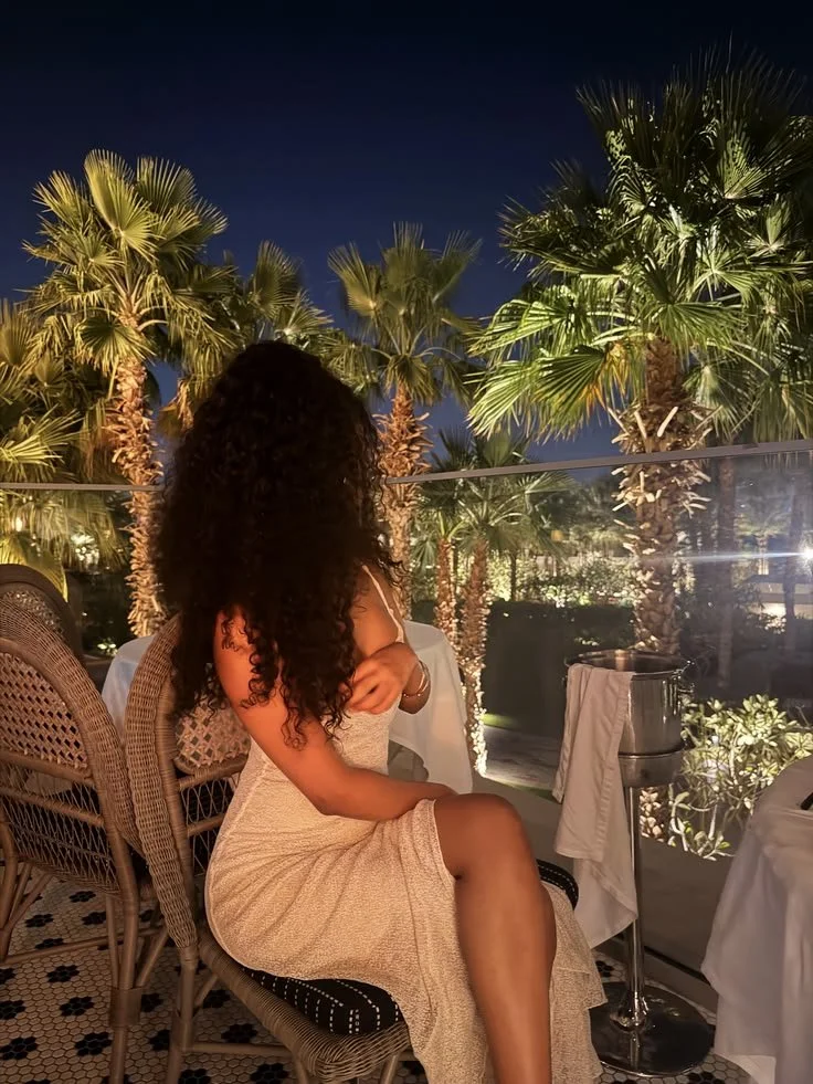 A woman with long curly hair sitting at an outdoor restaurant or patio at night, surrounded by illuminated palm trees.