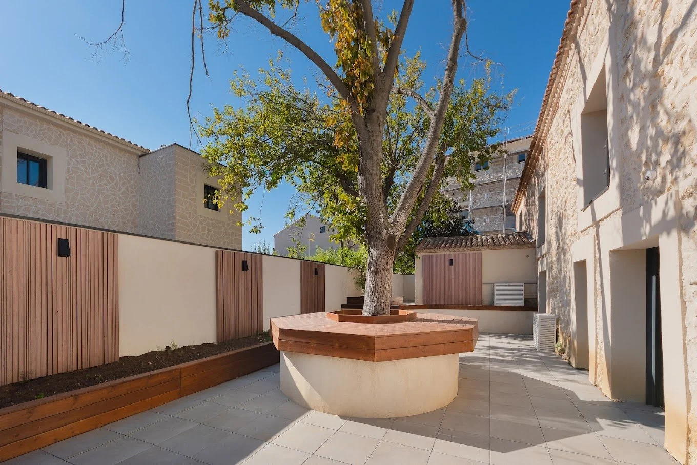 Aménagement extérieur d’une cour avec arbre central, assises en bois et murs en pierre.