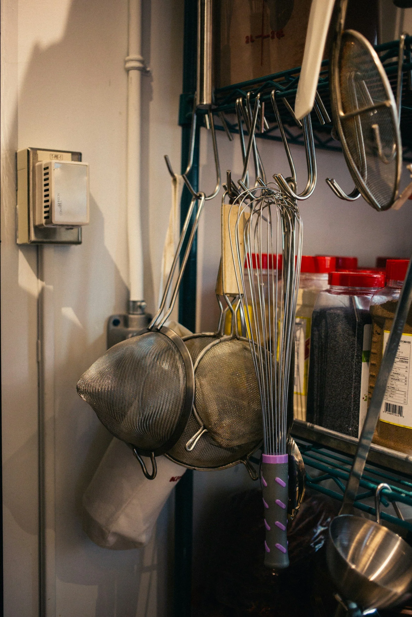Kitchen tools hanging on a rack, including metal strainers, metal skewers, and a pink-handled spatula, with jars and packaging in the background.