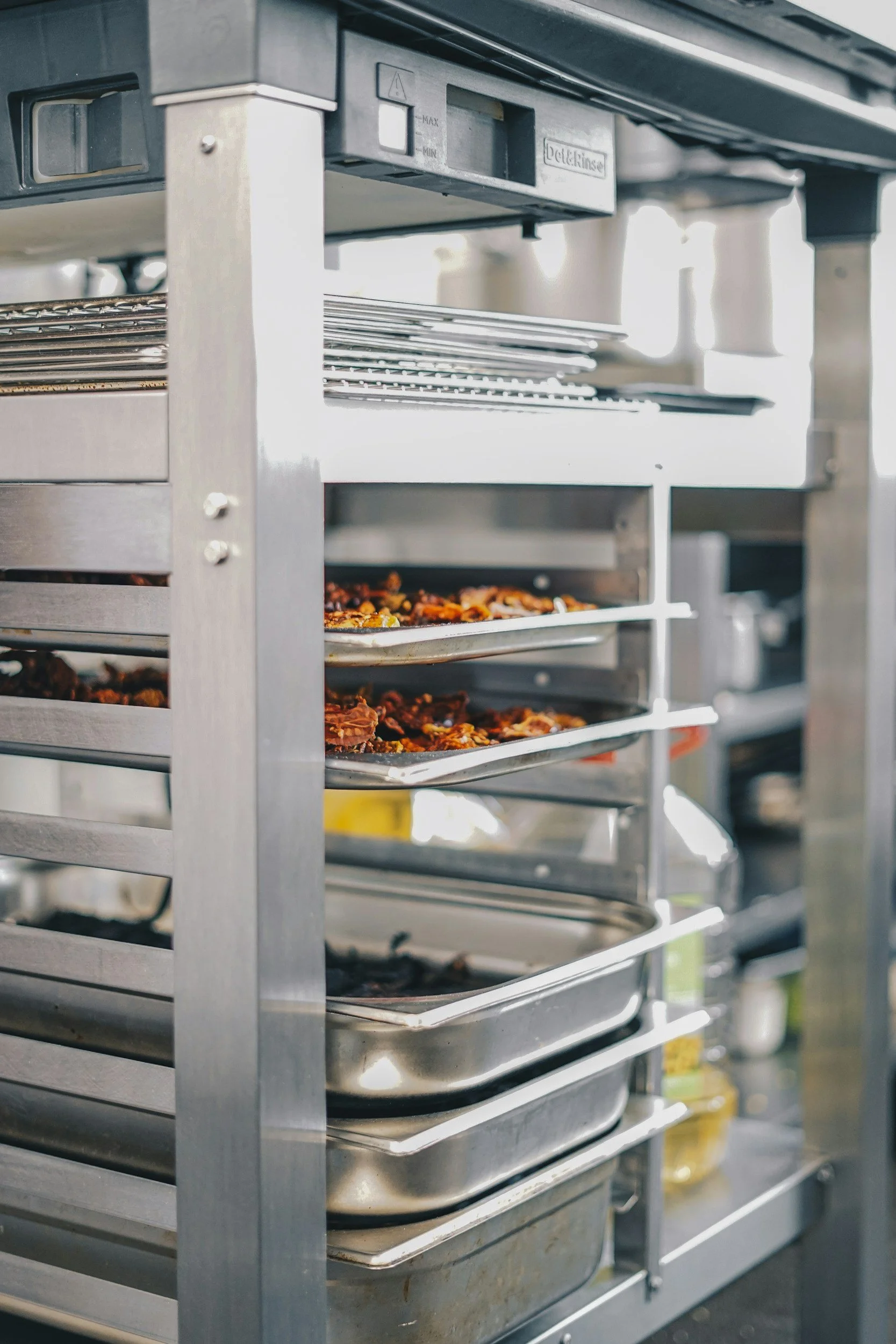 A stainless steel tray rack holding multiple oven trays with baked food in a commercial kitchen.
