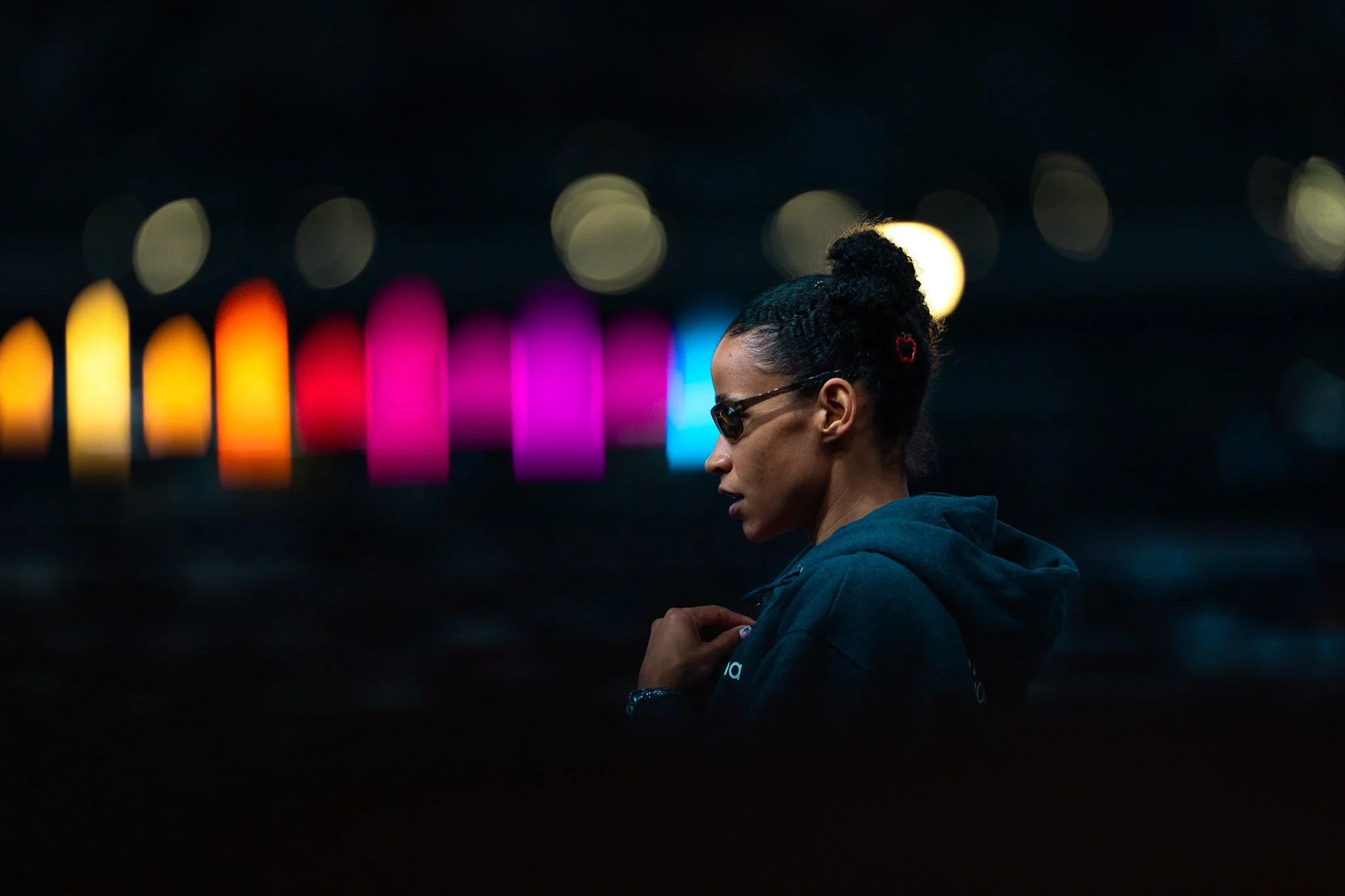 A woman with curly hair up in a bun, wearing sunglasses and a dark hoodie, stands in profile against a dark background with colorful blurred lights creating a bokeh effect.