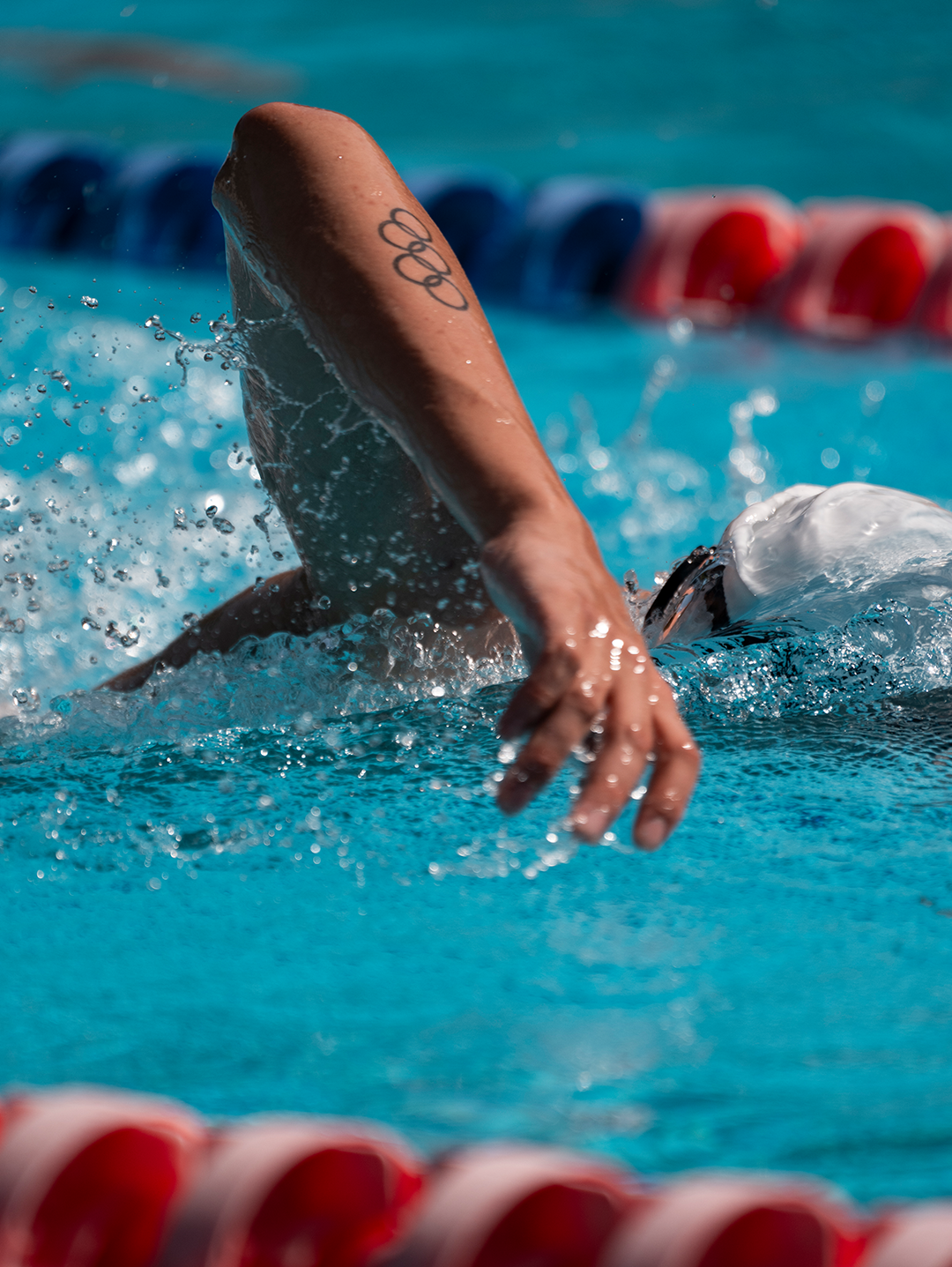 Close-up of a swimmer's arm and hand emerging from the water during a swimming race, with a tattoo on the upper arm, in a pool with lane markers in the background.