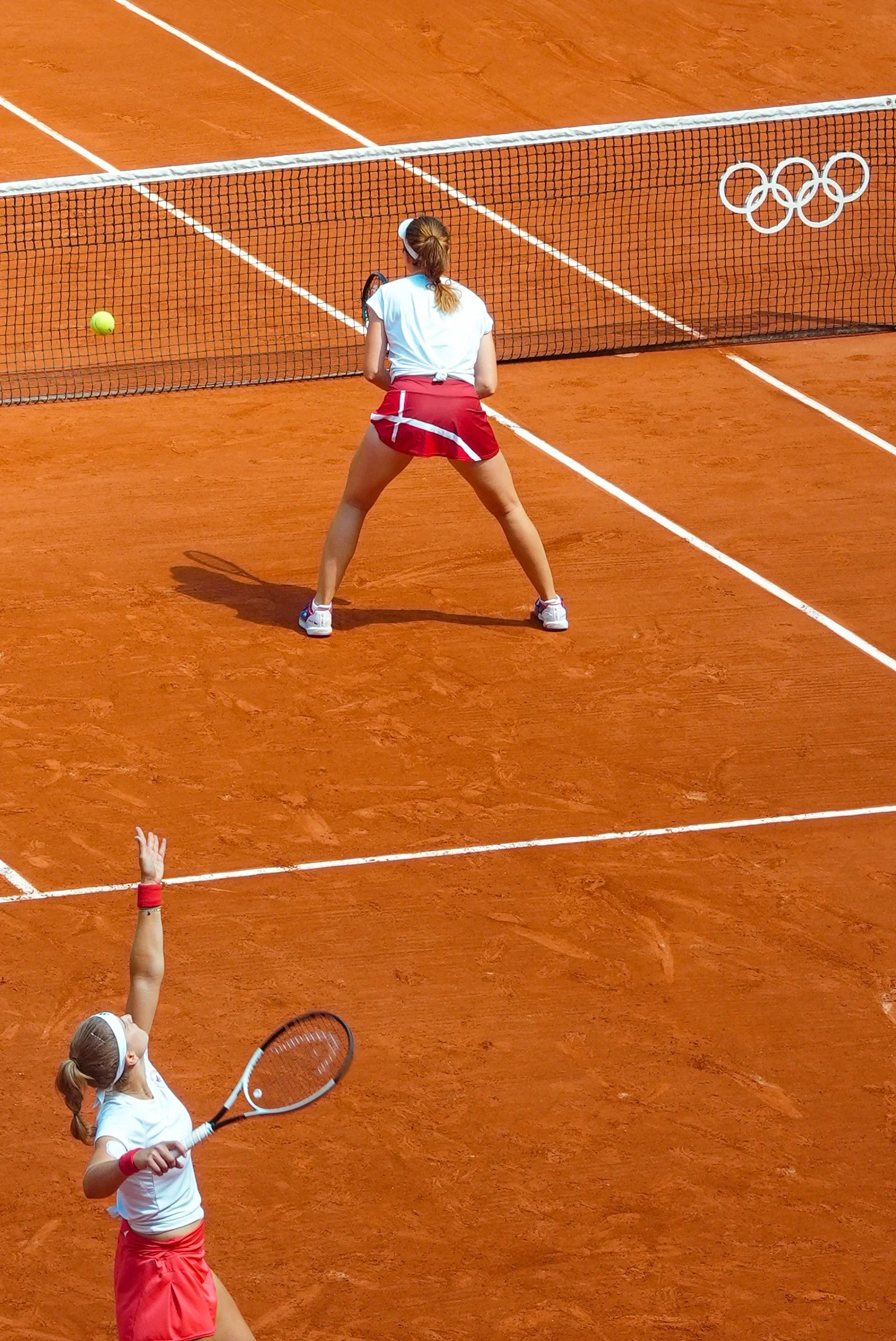 Tennis players during a match on a clay court, with one female preparing to hit the ball and another reaching to return, the Olympic rings are visible on the court.