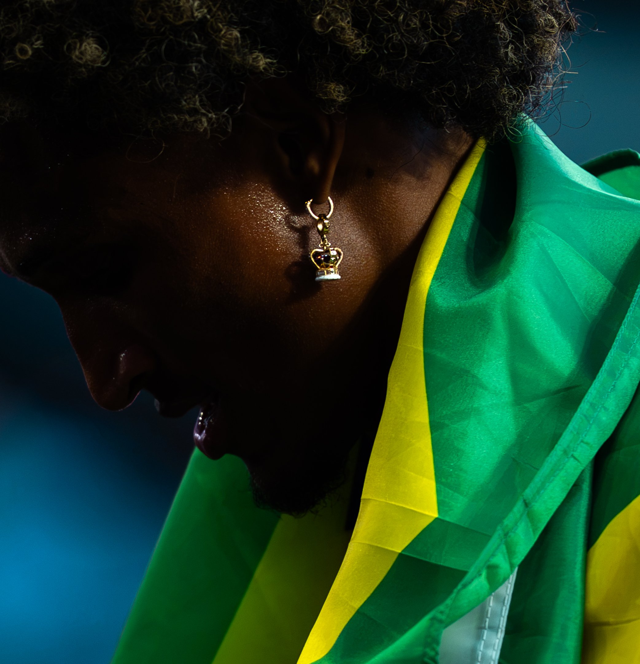 Close-up of a woman's face with dark skin, curly hair, and a nose ring, looking downward, wearing a green and yellow garment, against a dark background.