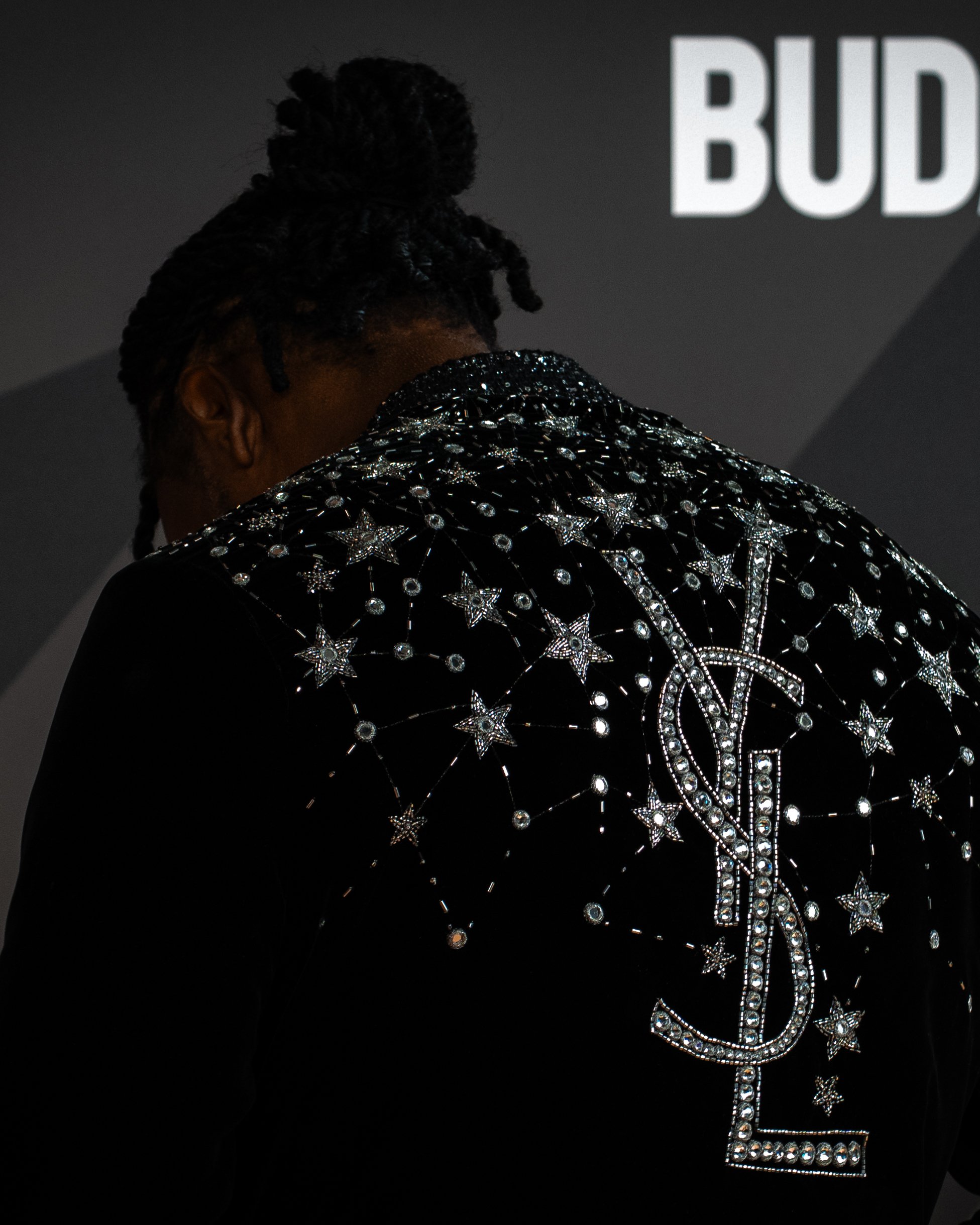 Close-up of a person wearing a black shirt embroidered with silver stars and other celestial designs, with a dark background partially showing the text 'BUD'.
