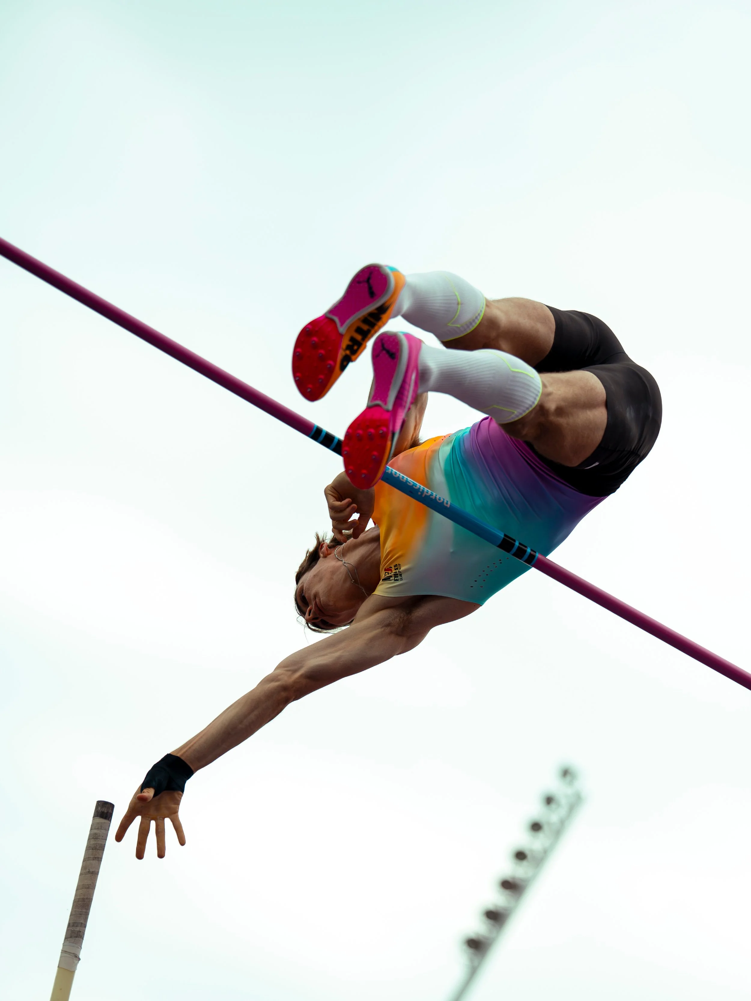 An athlete in colorful sportswear and white socks performs a pole vault jump, clearing the bar against a cloudy sky.